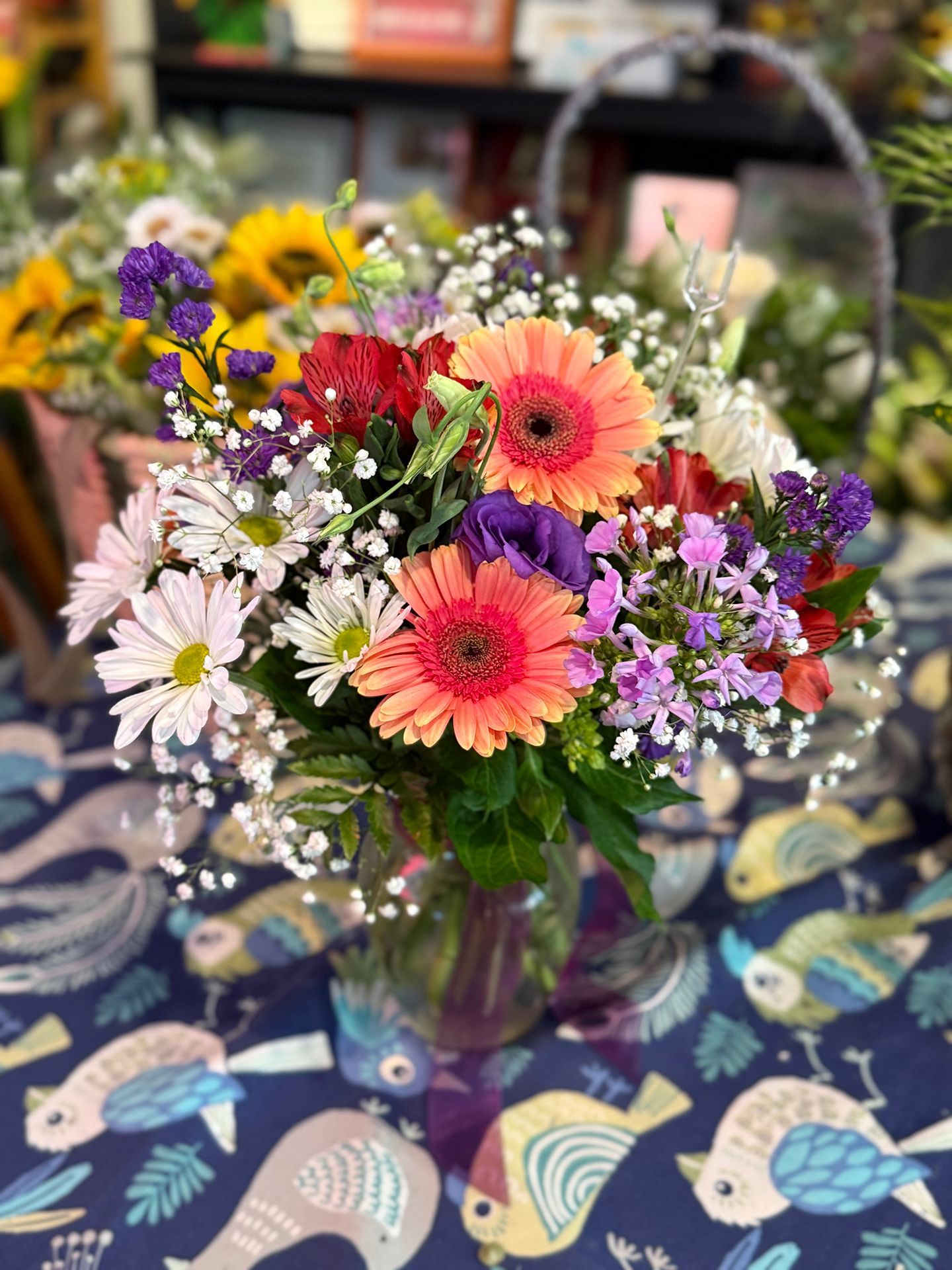 Close-up of a colorful flower arrangement in a glass vase, featuring gerbera daisies, daisies, and other flowers, on a patterned tablecloth.