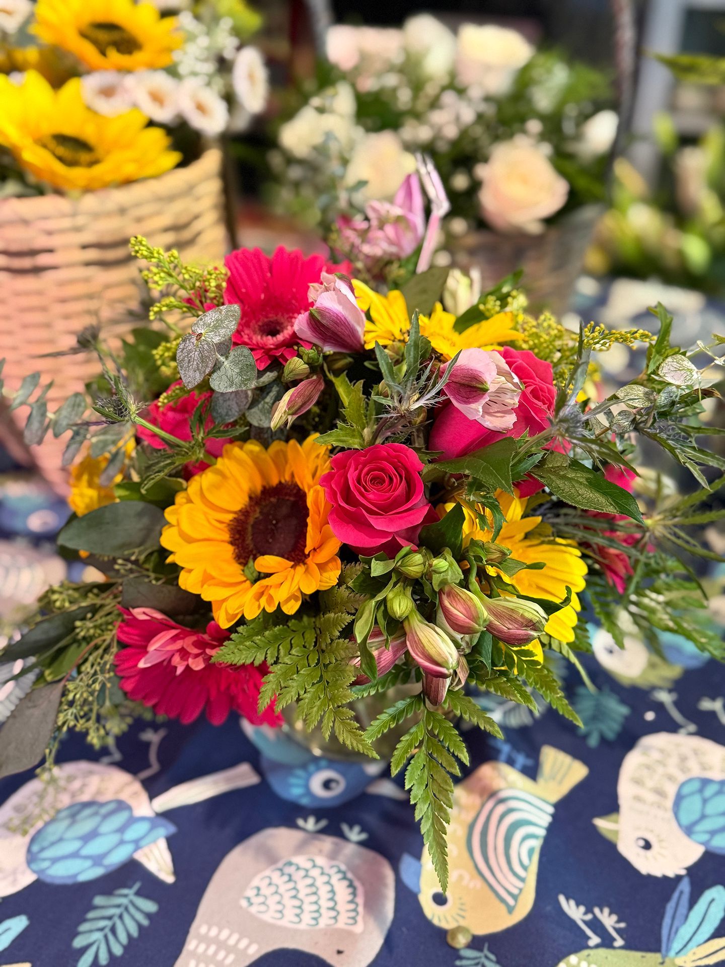 Close-up of a vibrant floral arrangement in a blue vase featuring sunflowers, red roses, and green foliage on a patterned surface.