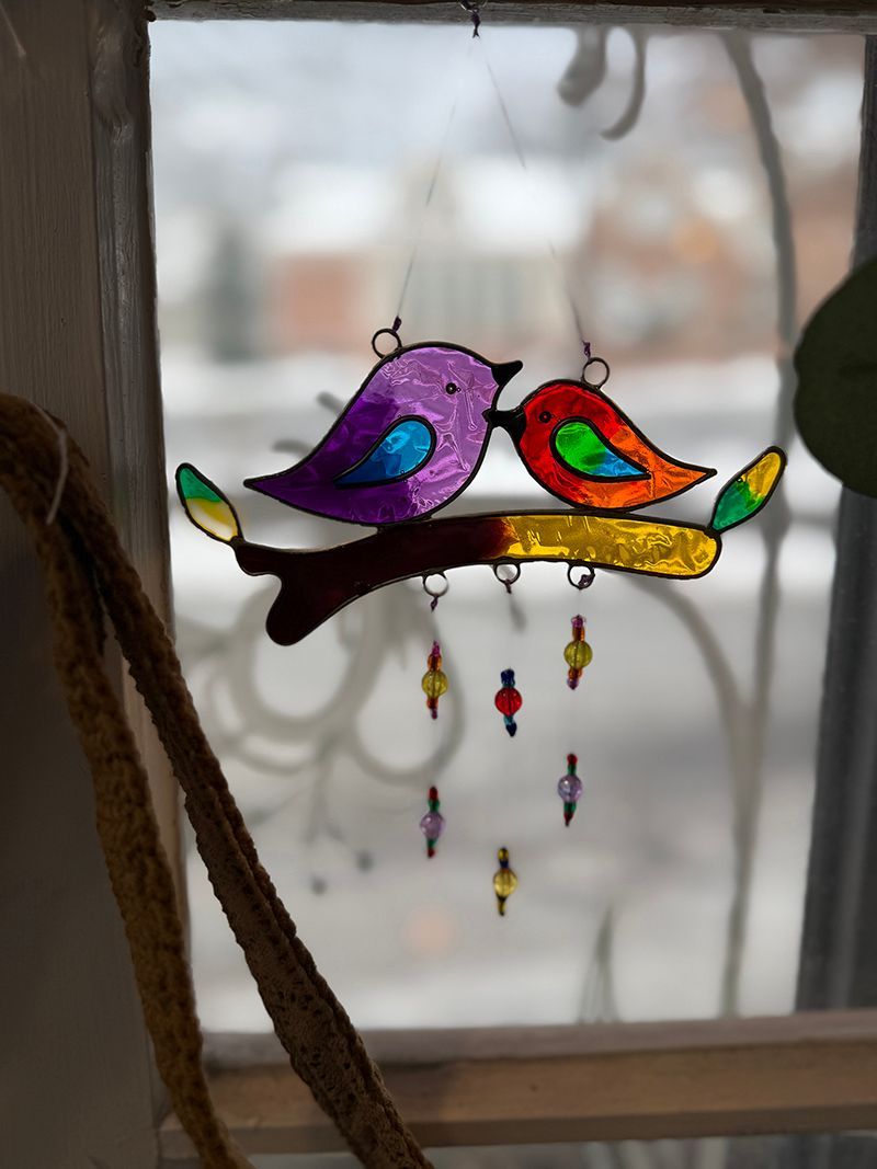 Stained glass birds kissing on a branch, hanging in a window. Purple and red birds with colorful details.