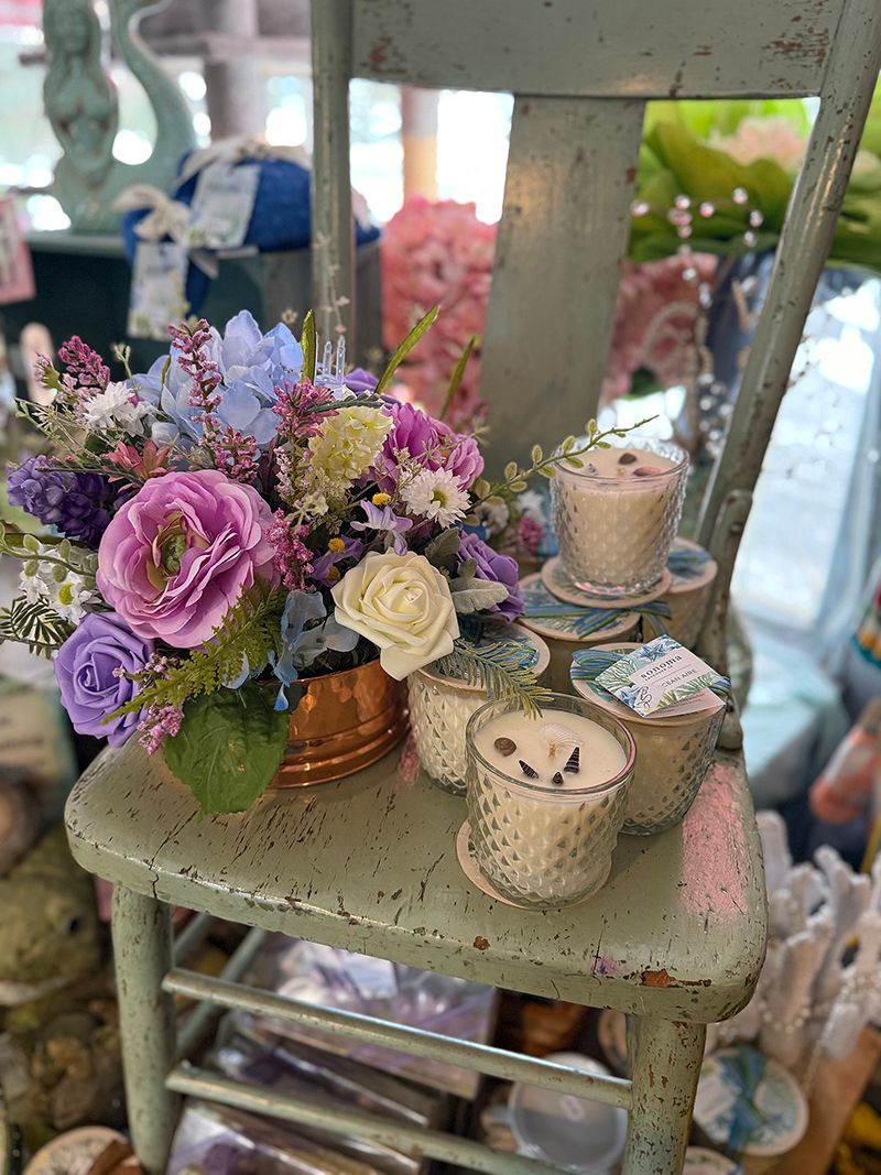 A floral arrangement in shades of purple and blue on an antique green chair next to candles with shells.