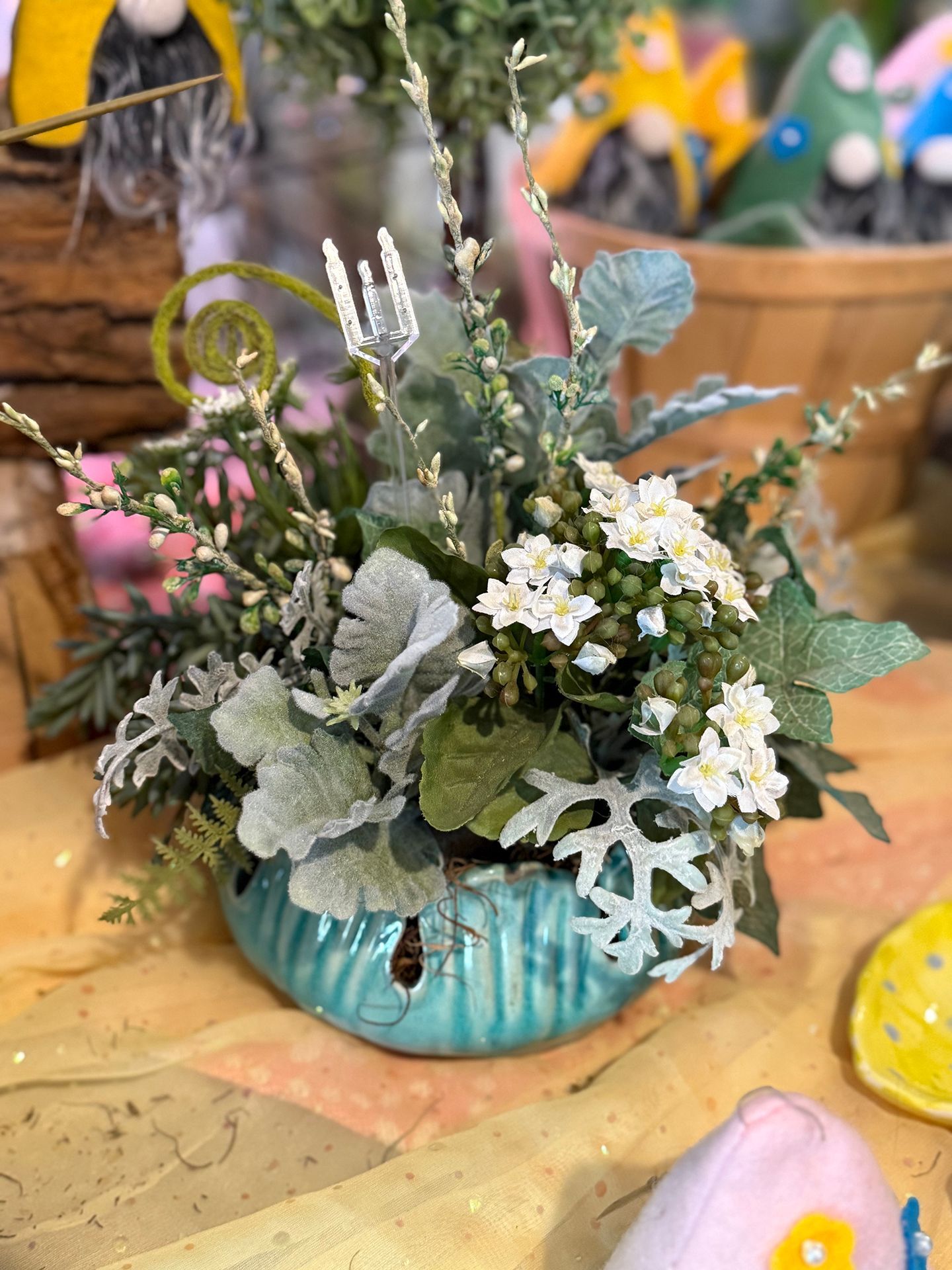 A small, round, turquoise ceramic planter with an arrangement of white and green flowers and foliage. The setting is a shop.