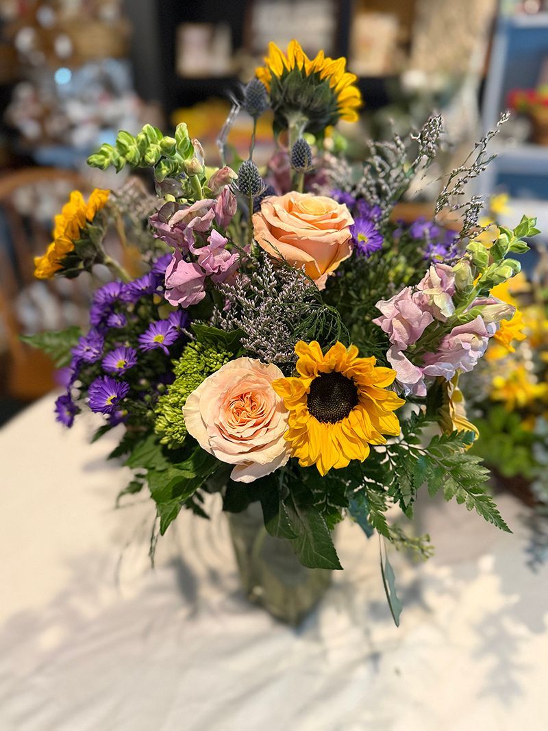 Vibrant bouquet with sunflowers, roses, and purple flowers in a glass vase on a white table.