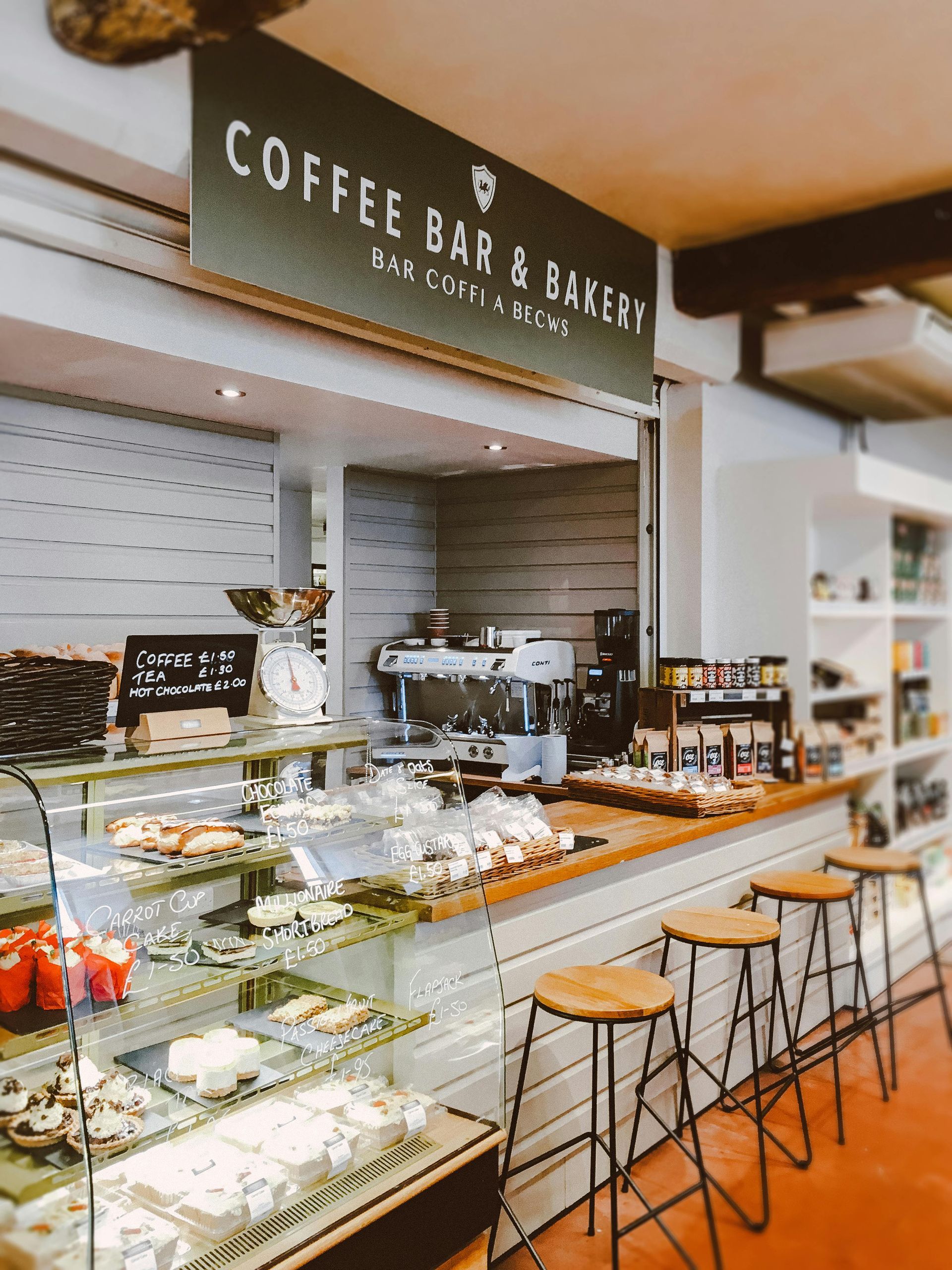 Interior de cafetería y panadería con pasteles en exhibición, máquina de café expreso y asientos.