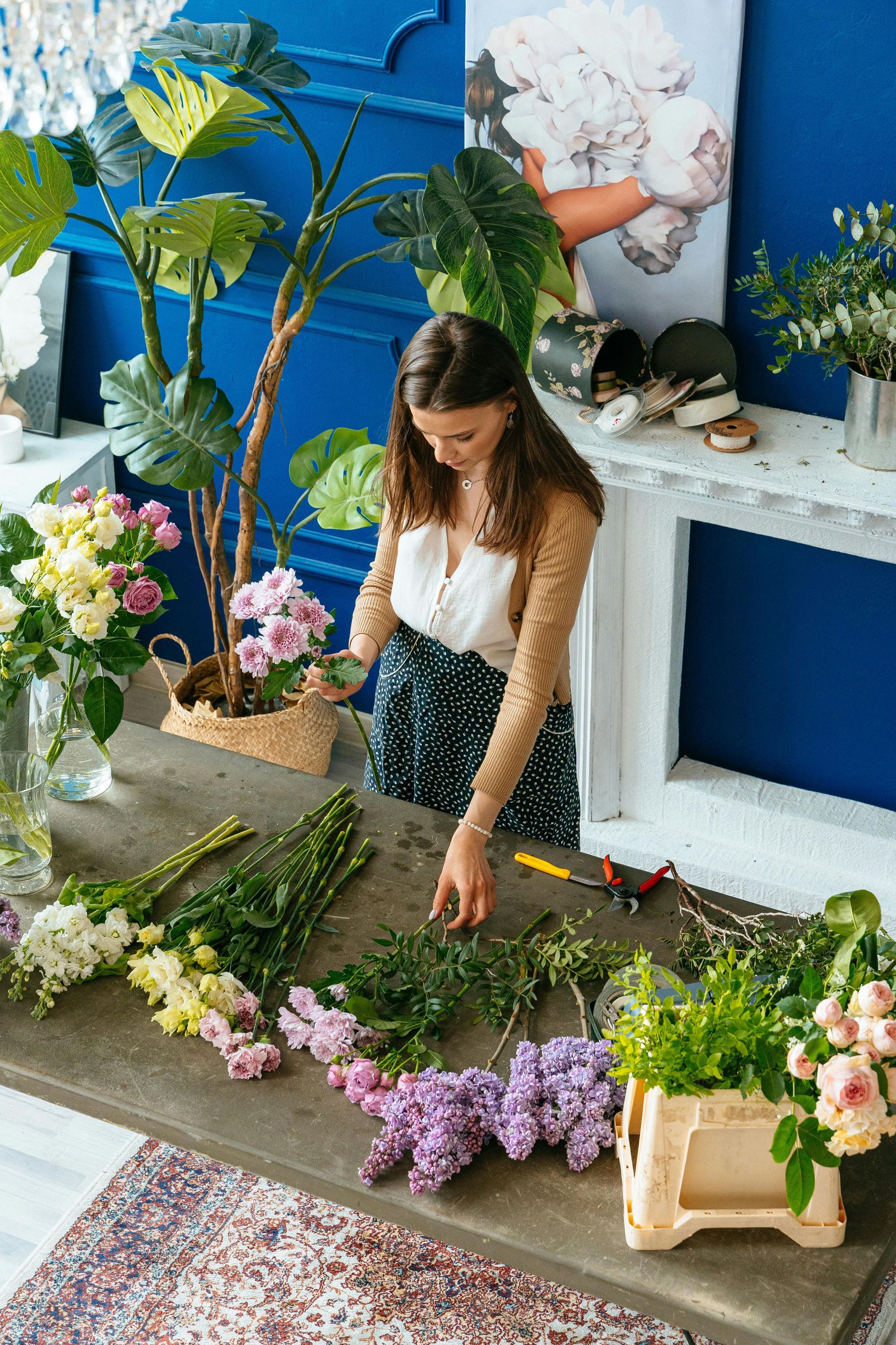 Mujer arreglando flores en una mesa con vegetación. Pared azul, blusa blanca y falda negra de lunares.