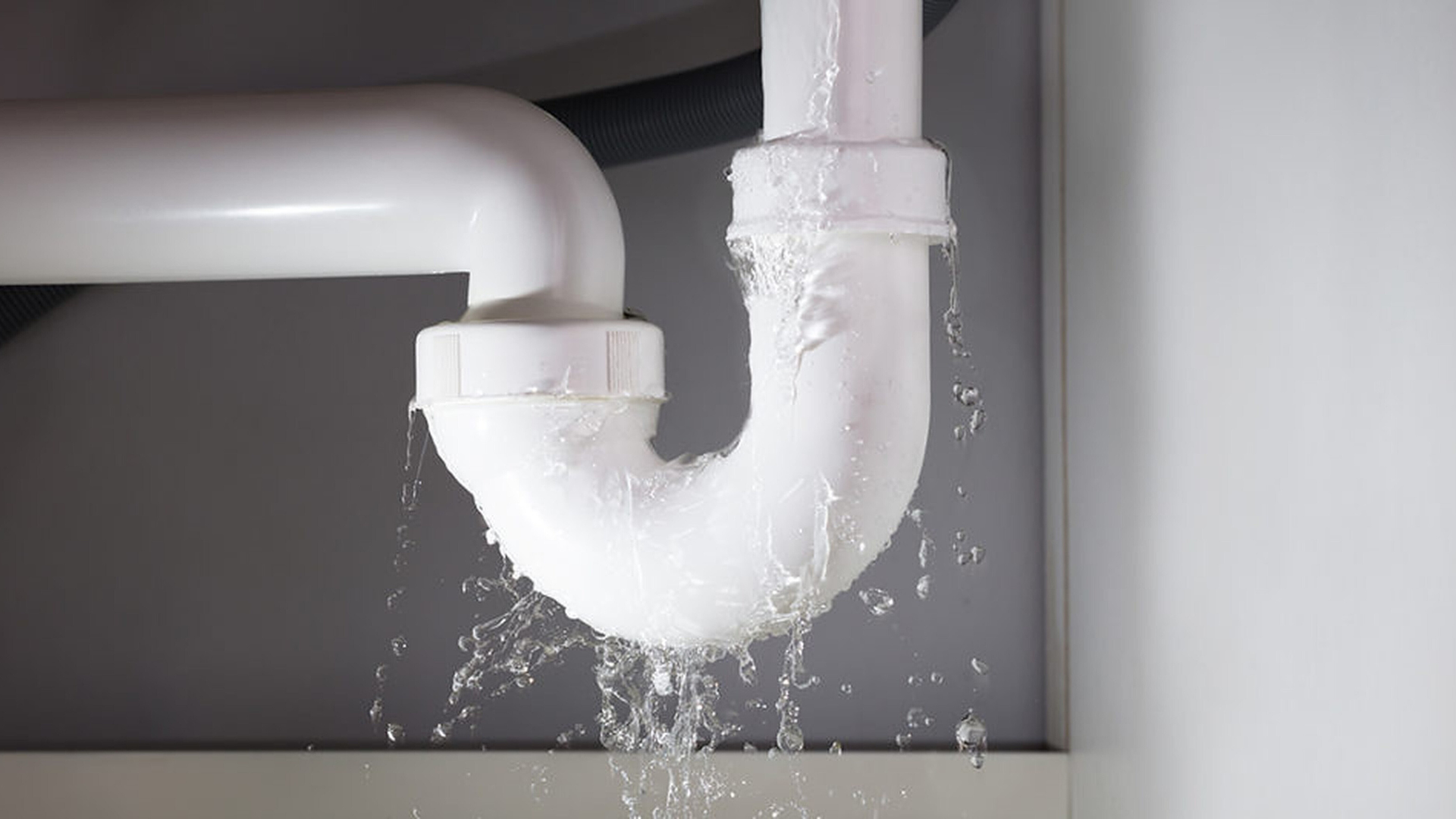 Leaking white PVC plumbing pipes in a gray cabinet, with water dripping down.