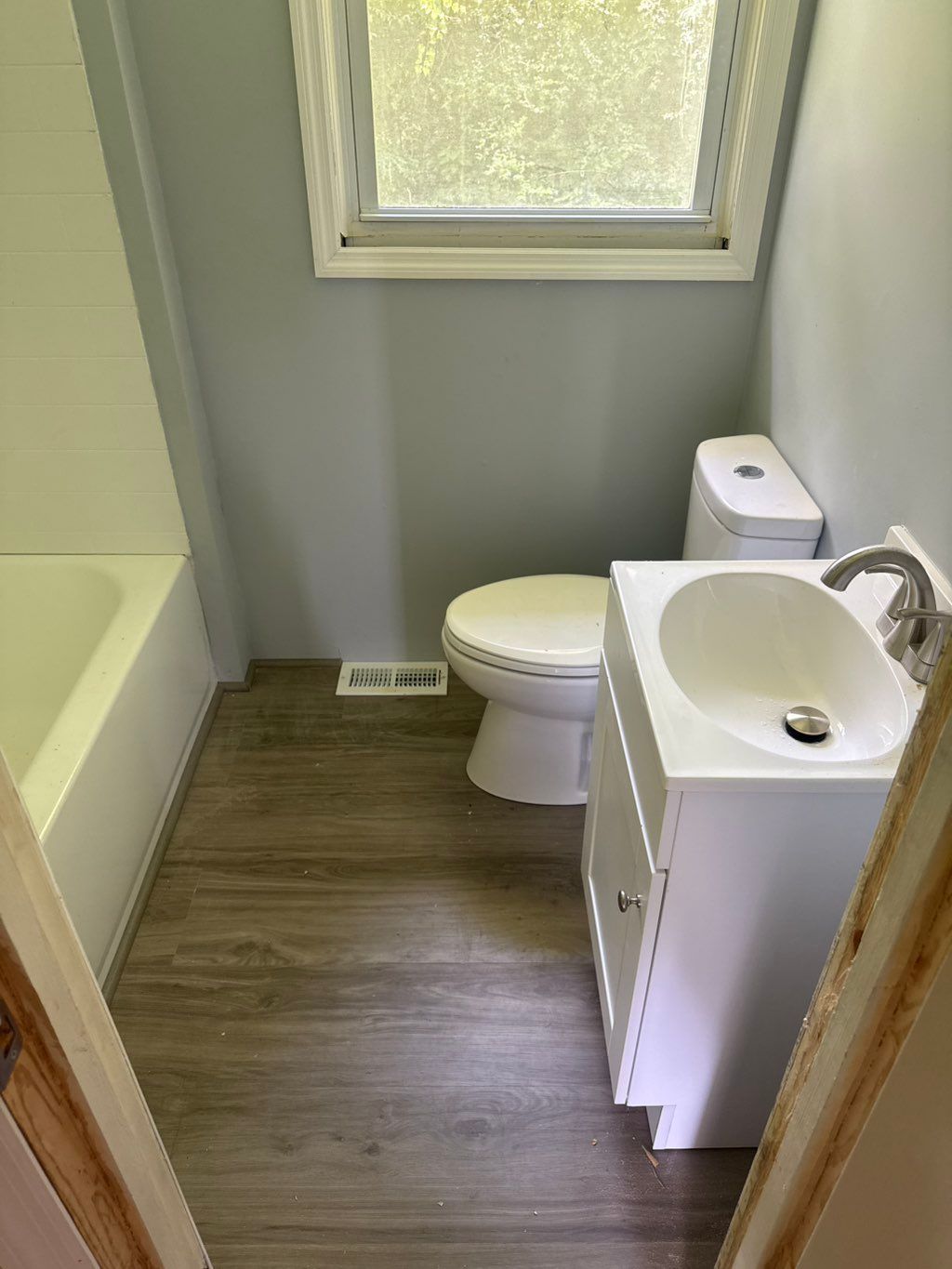 Small bathroom with a toilet, sink, and tub, painted light blue with a wood-look floor.