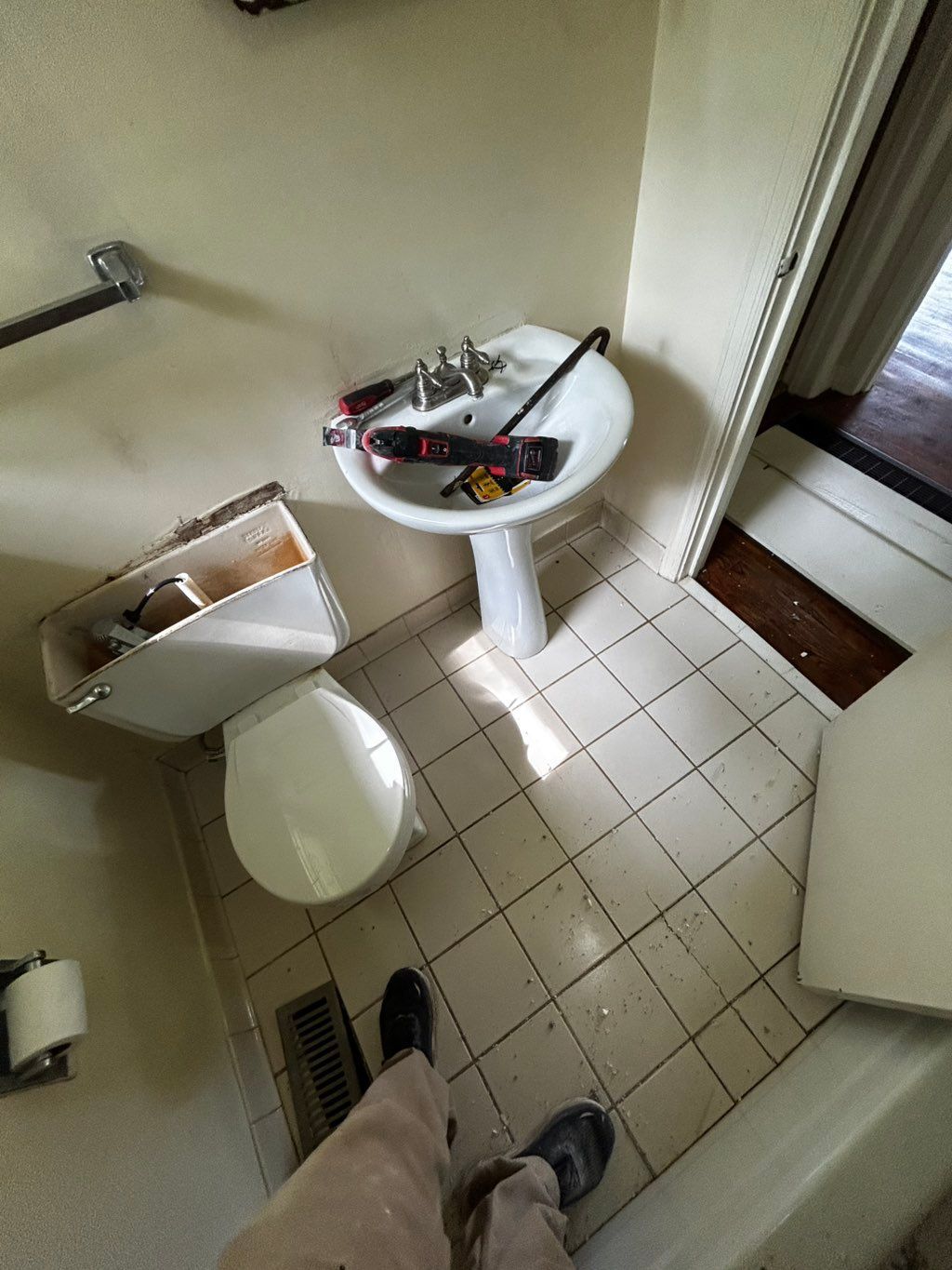 Bathroom with a pedestal sink, toilet, and tools on the sink; a person's feet are visible.