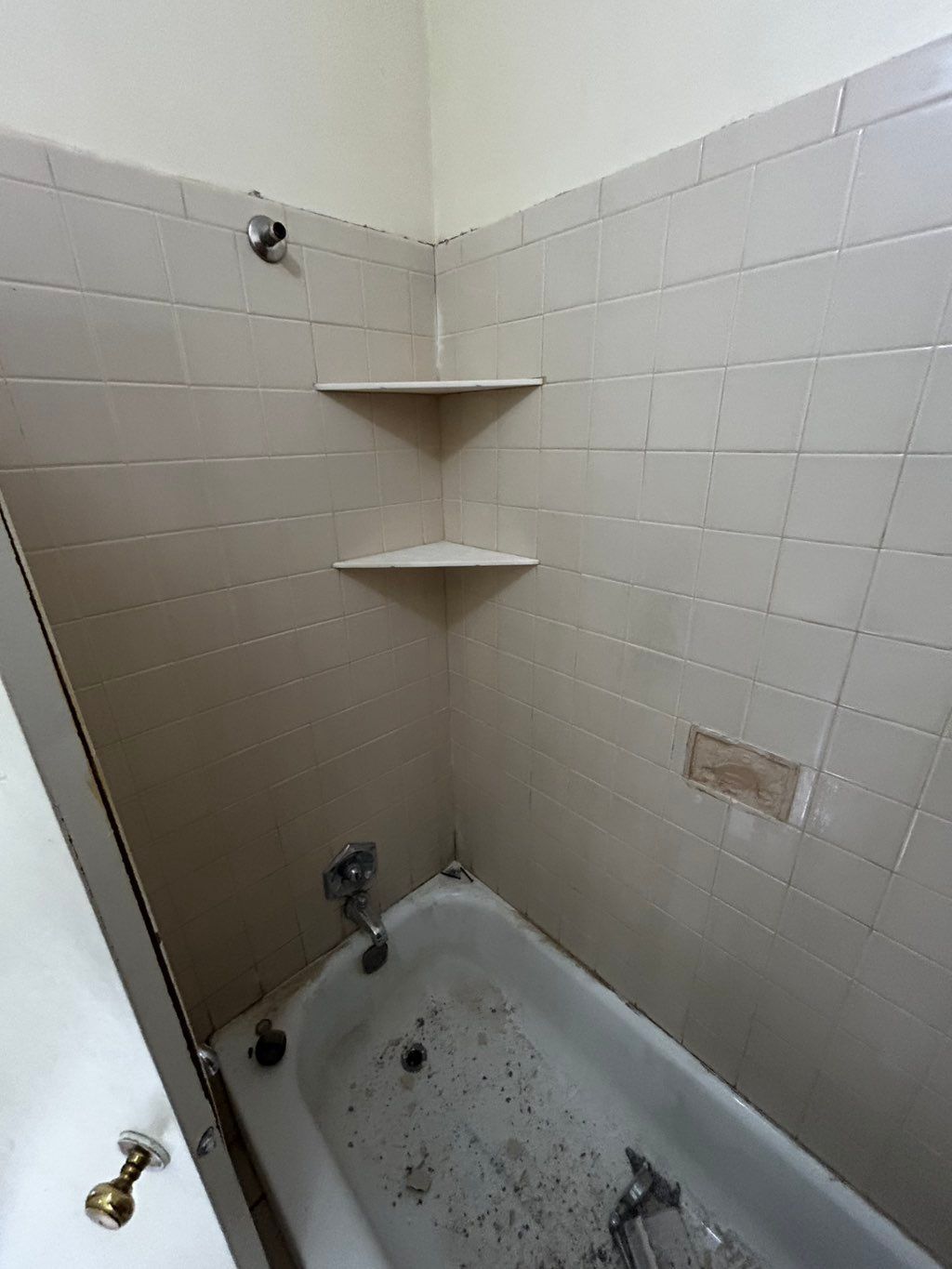 Corner shower with two shelves, white tiles, and a tub, in a bathroom.