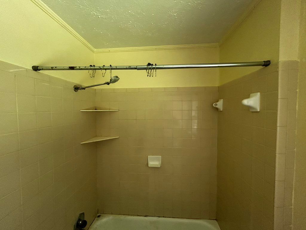 A small beige-tiled bathroom with a shower rod, shelves, and fixtures.