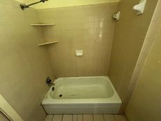 A small, tiled bathroom with a white bathtub, two shelves, and a shower head.