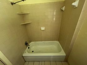 A small, tiled bathroom with a white bathtub, two shelves, and a shower head.