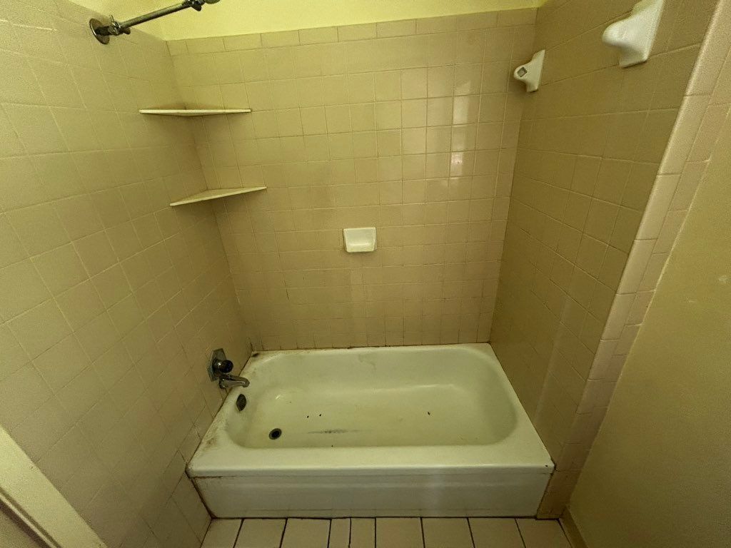 Small, beige-tiled bathroom with a bathtub, shelves, and showerhead.