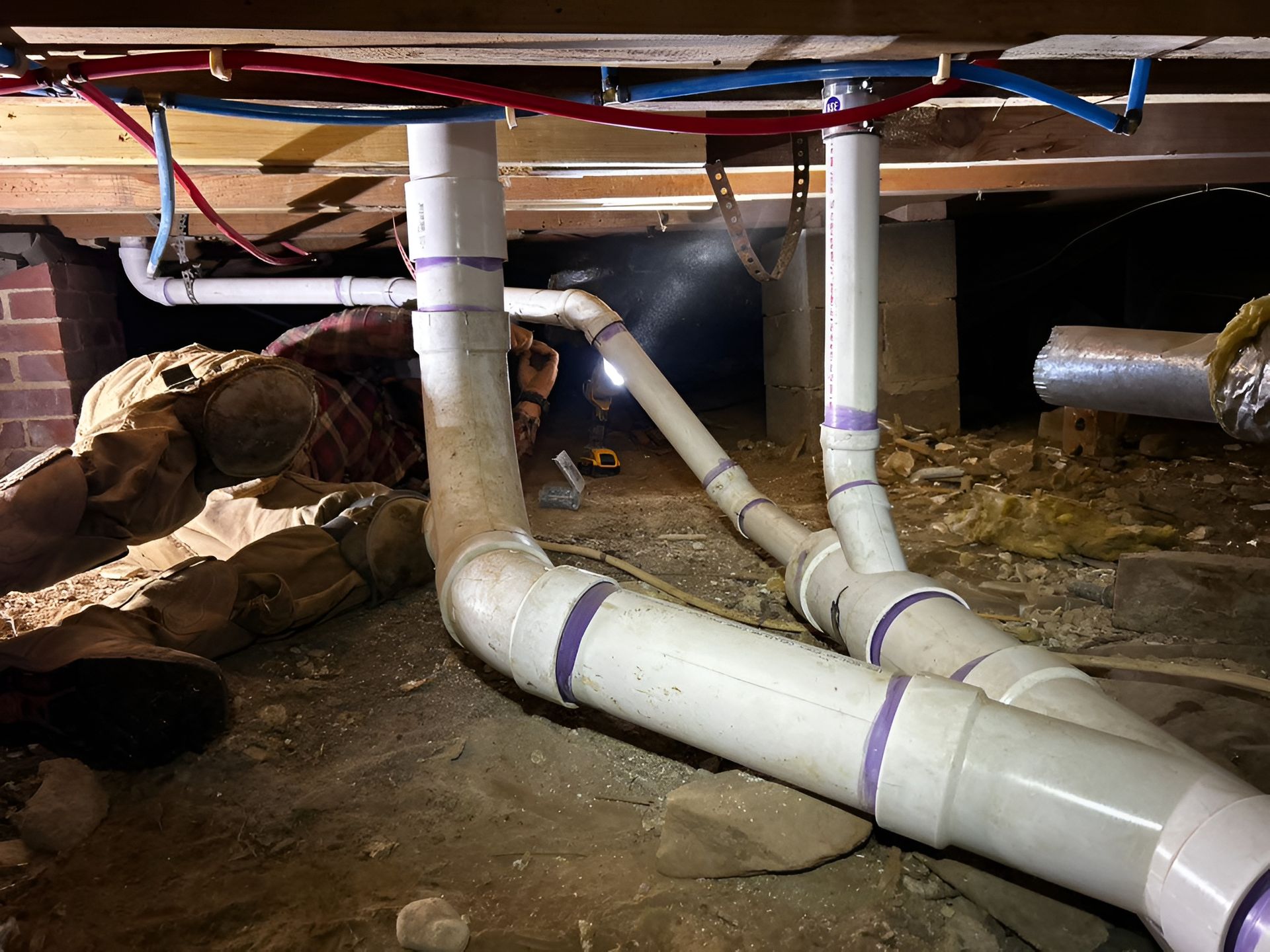 Plumber working under a house, installing PVC pipes. The area is dimly lit.