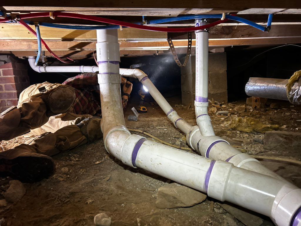Plumber under house fixing pipes. White PVC pipes, red/blue water lines, dark underbelly, and exposed wooden beams.