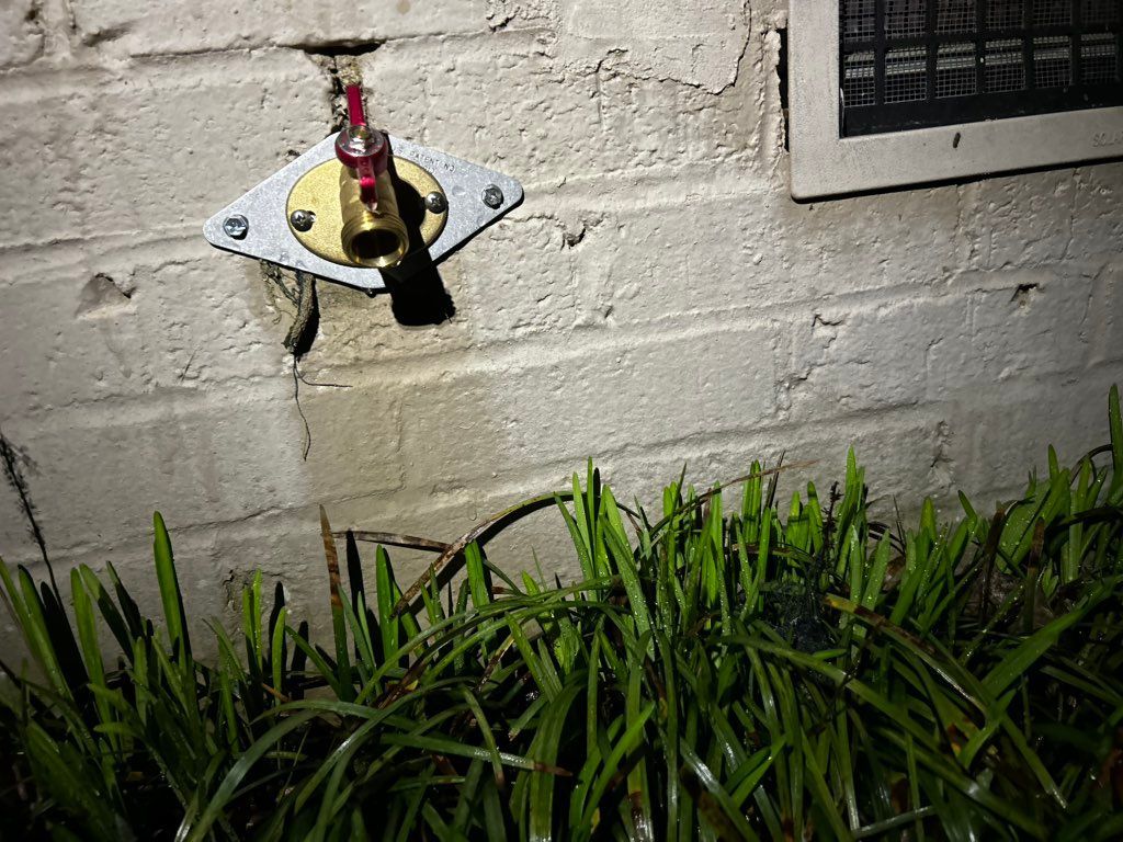 Outdoor water spigot attached to a brick wall, above green grass.