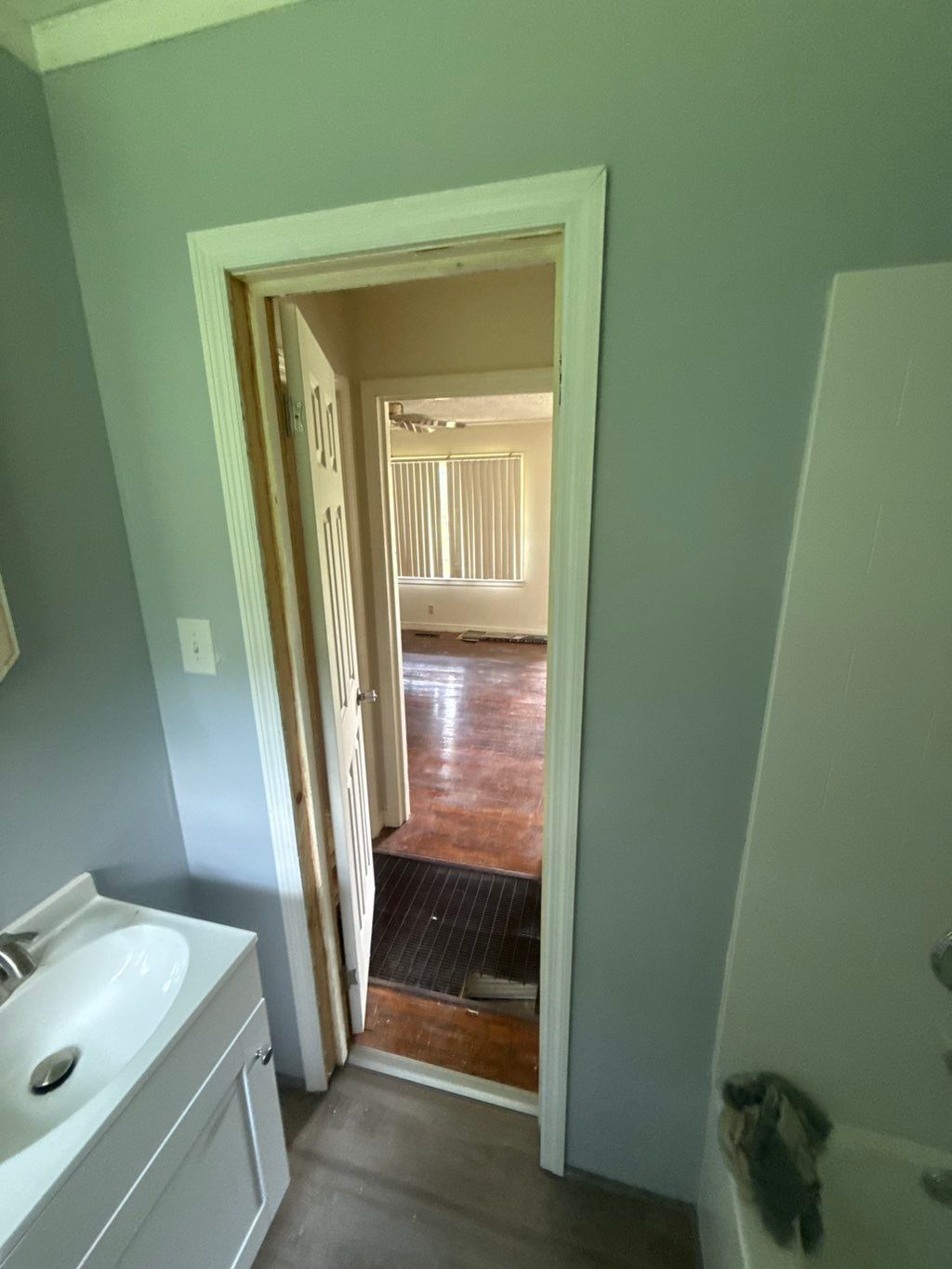 Open doorway into a room, viewed from a bathroom with a sink, pale green walls, and brown flooring.