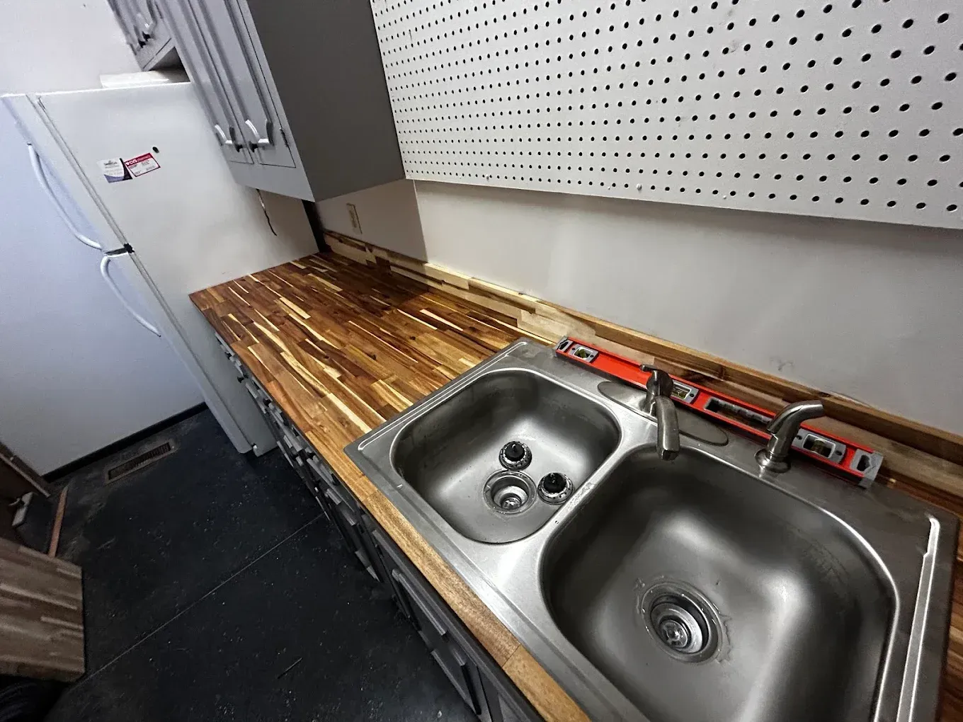 A kitchen with a stainless steel sink, wooden countertop, refrigerator, and pegboard wall.