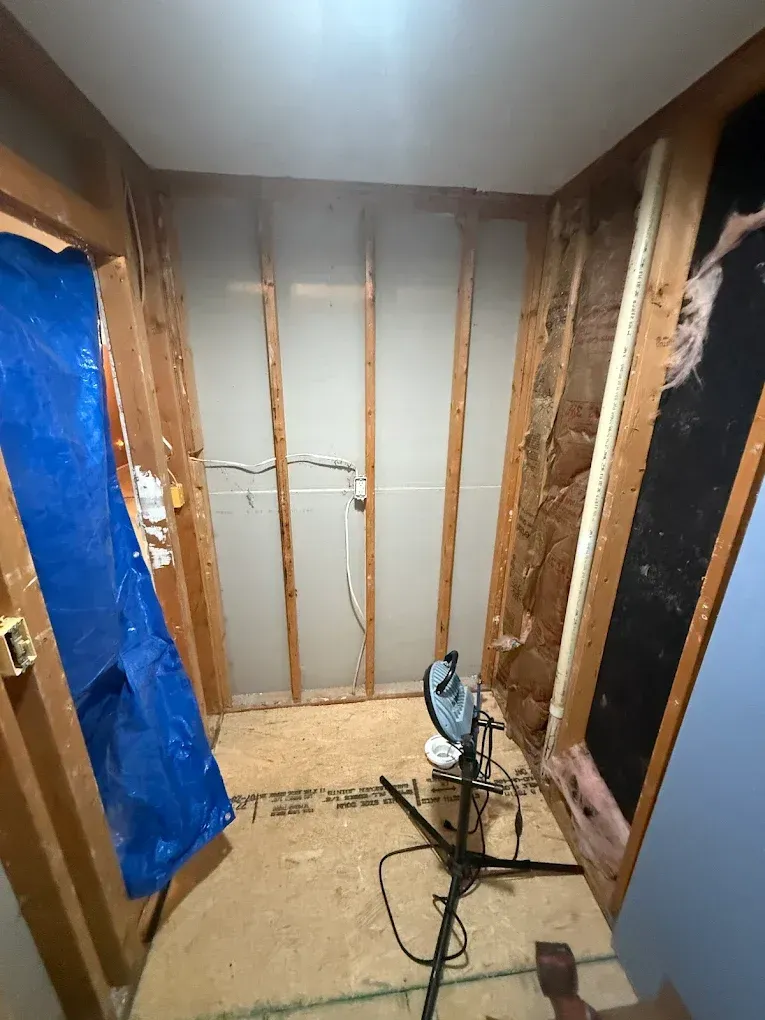 Interior view of a small room under construction, with exposed wooden framing and drywall. A blue tarp hangs on the left.