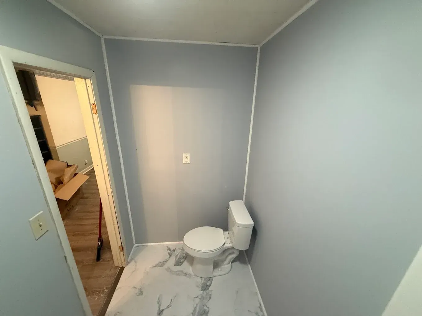 Small, blue-painted bathroom with white trim. A toilet sits on marbled flooring. Open doorway to a room.
