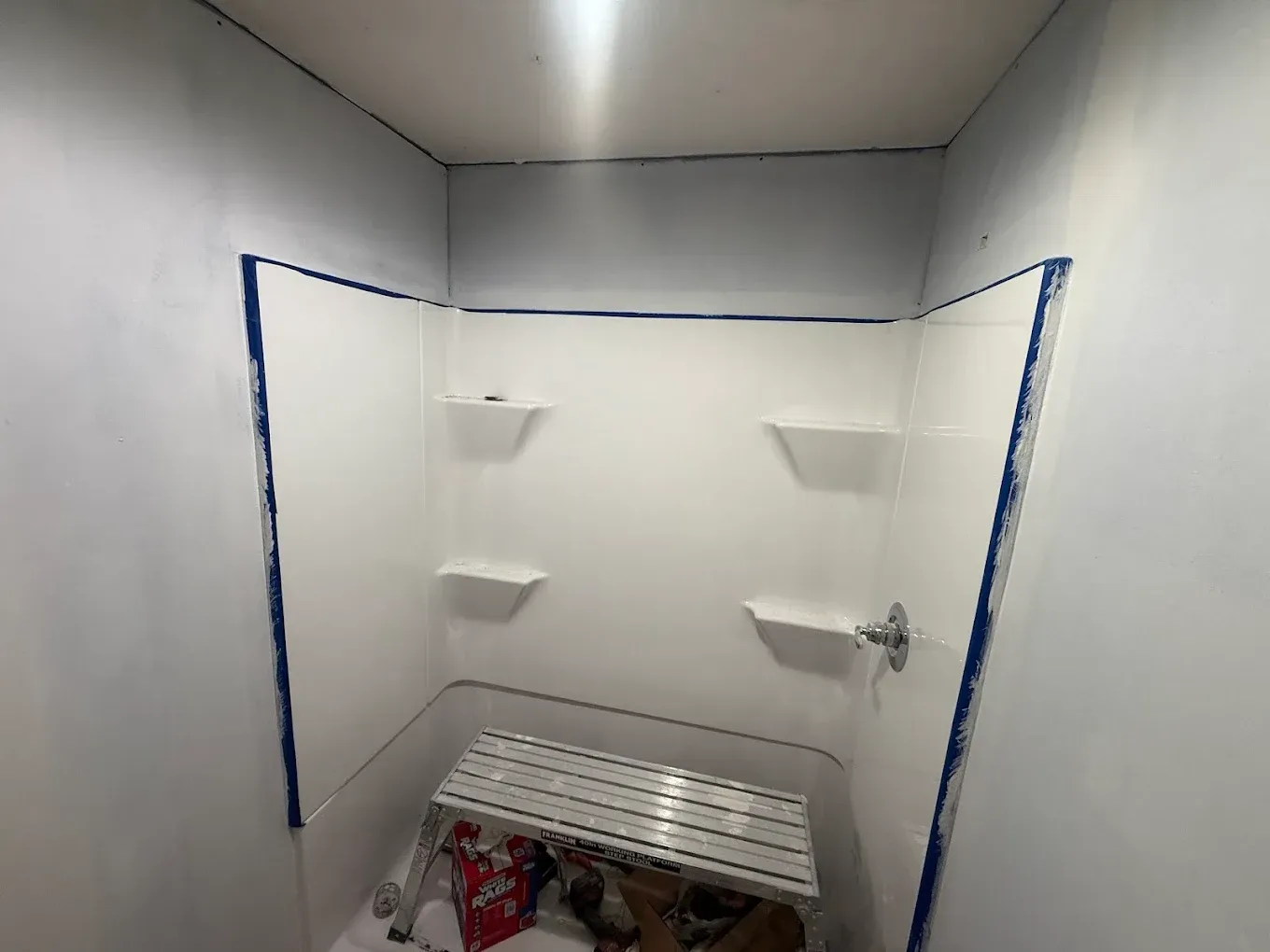 White shower stall interior with blue tape along edges, small shelves, and a step stool.