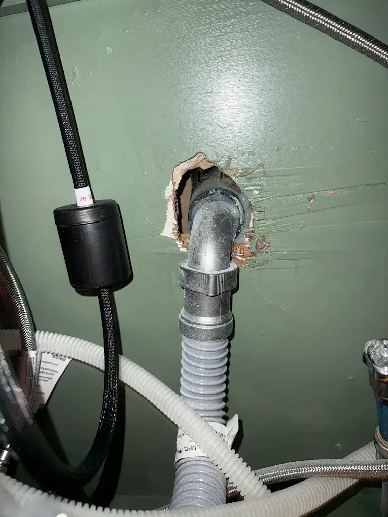 Plumbing under a green sink: drainpipe and a black cord. Hole in the wall.