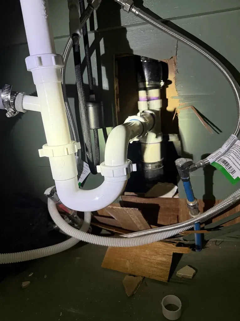 White plumbing pipes and fittings under a sink against a green wall, including a P-trap, drain, and supply lines.