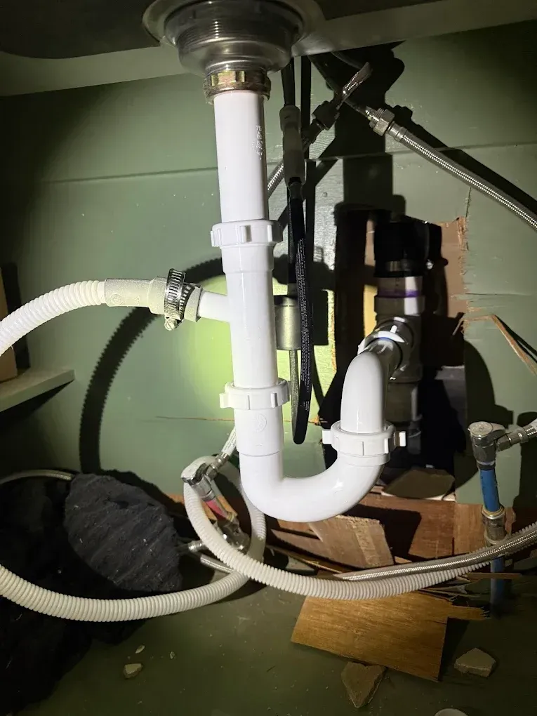 White plastic plumbing under a sink. Pipes and p-trap connected, with a black device and water lines visible.