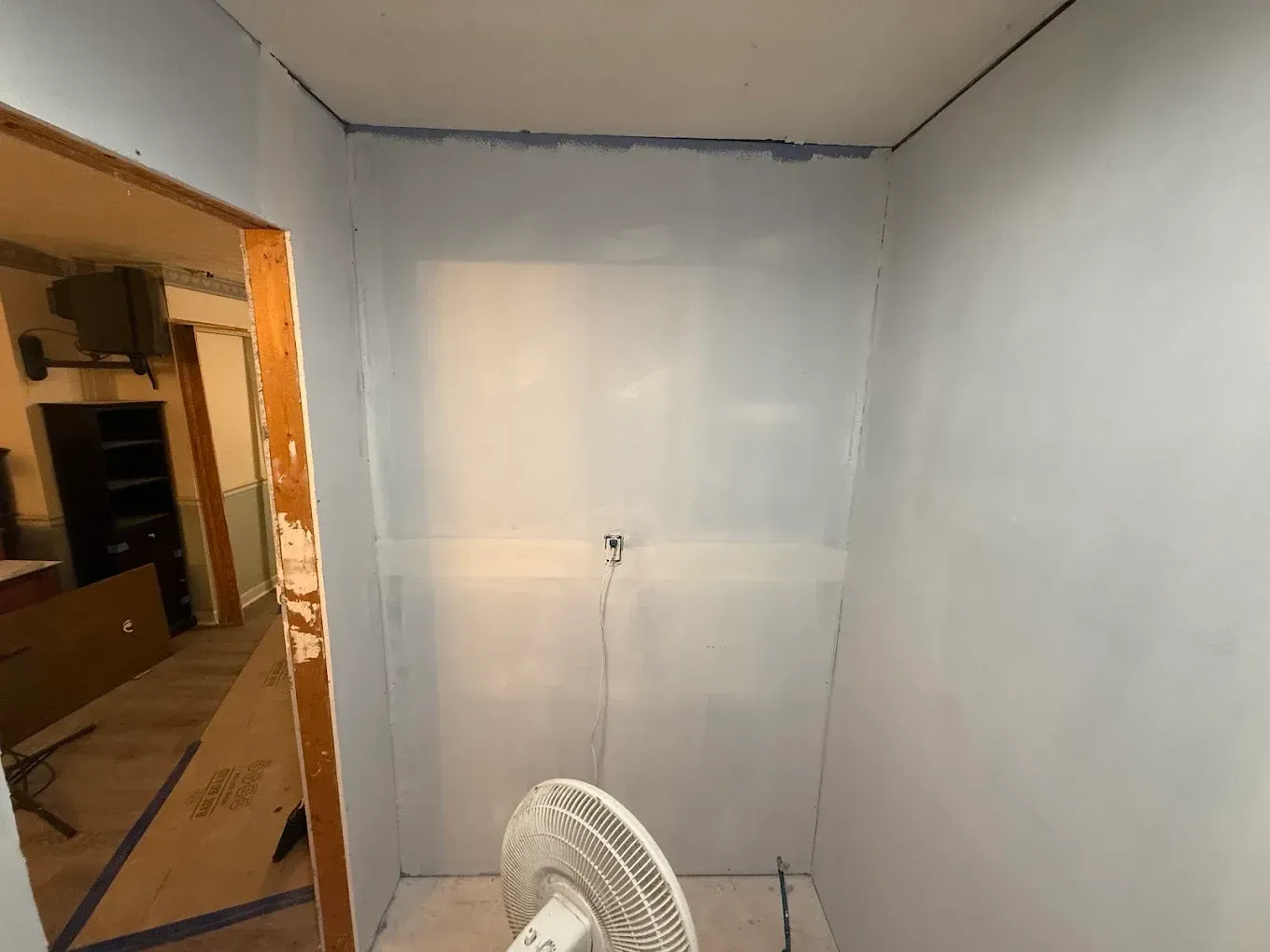 Interior of a small room with light blue walls, a white fan, and an open doorway to another room.