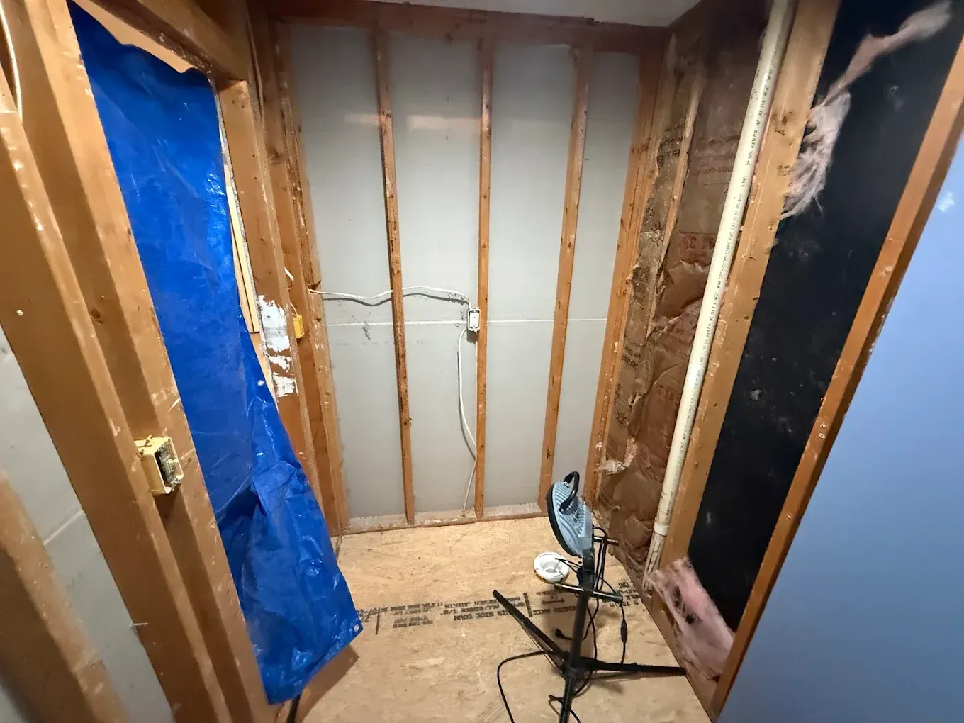Interior view of a small room undergoing renovation; exposed studs, insulation, tarp, and sawdust on the floor.