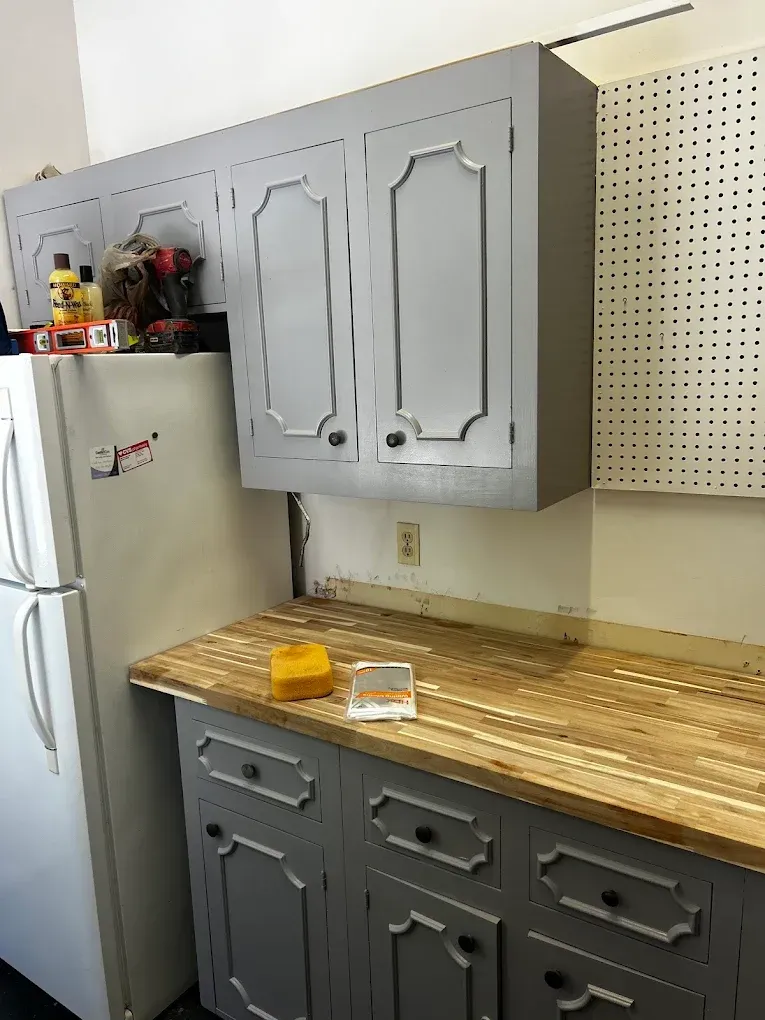 Gray kitchen cabinets, wooden countertop, refrigerator on the left, and pegboard wall on the right.