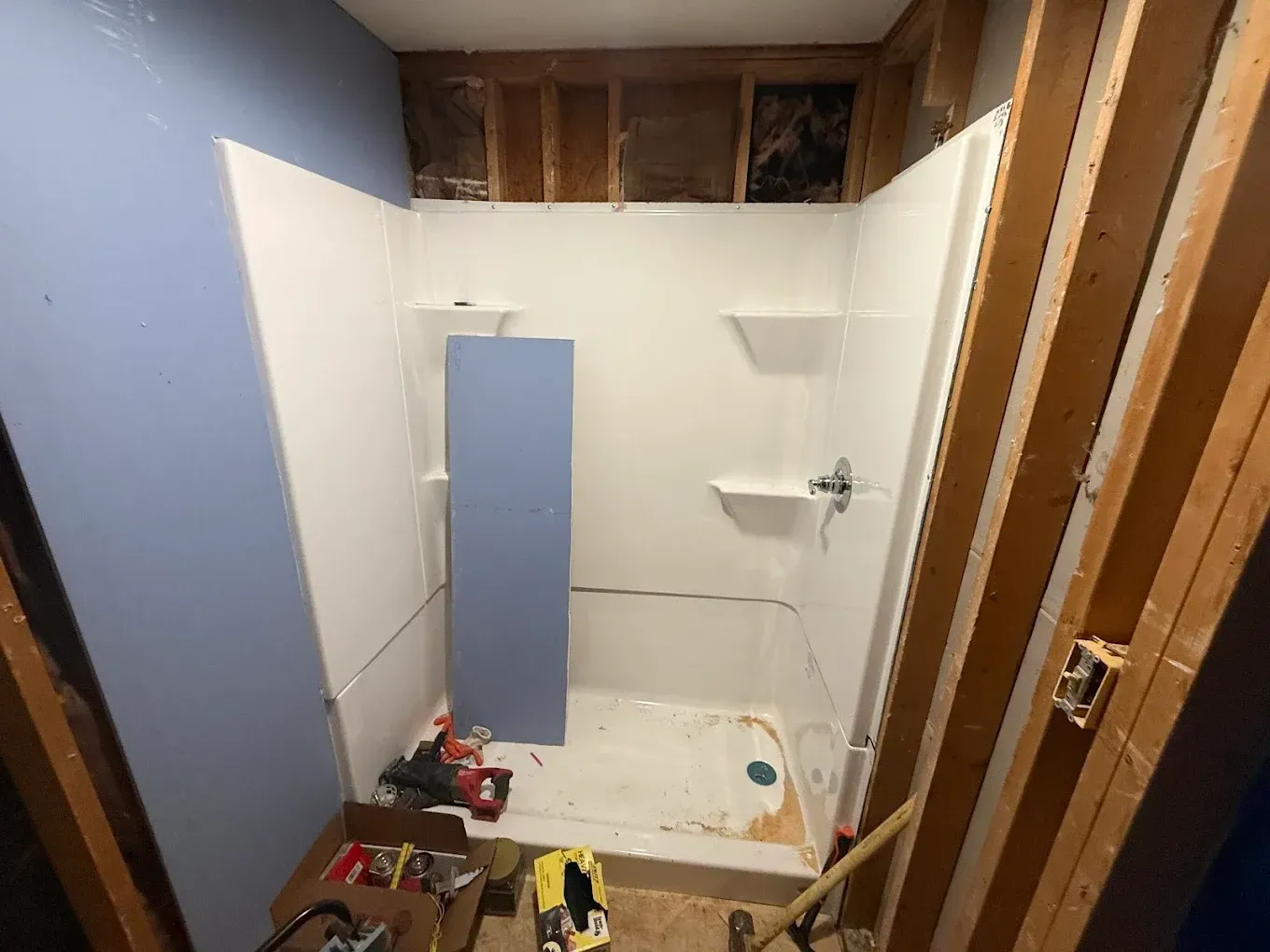 Bathroom renovation in progress, showing a white shower stall, exposed wooden framing, and blue/white walls.