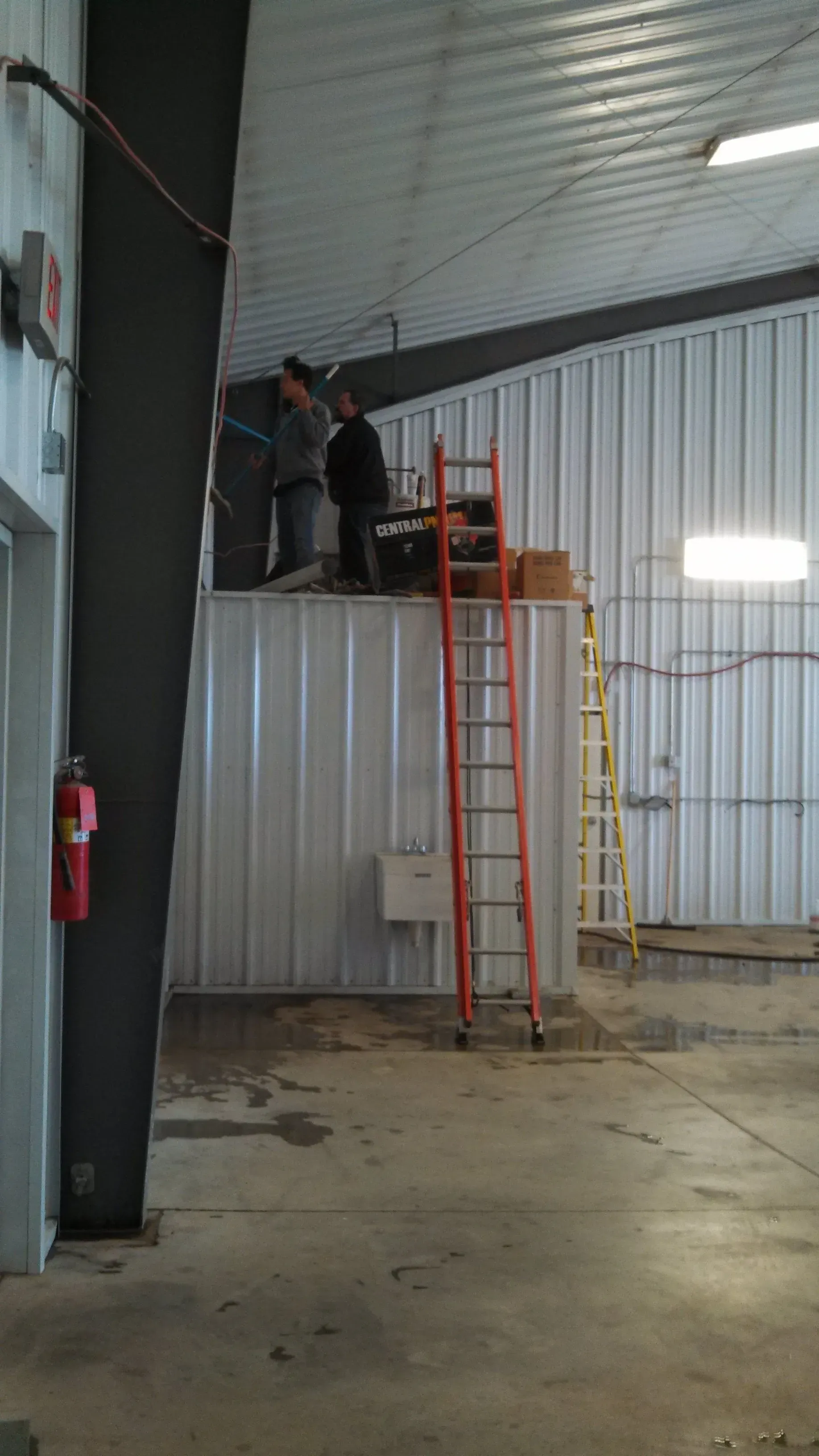 Two workers on a metal platform near a high ceiling, with a ladder and fire extinguisher visible.