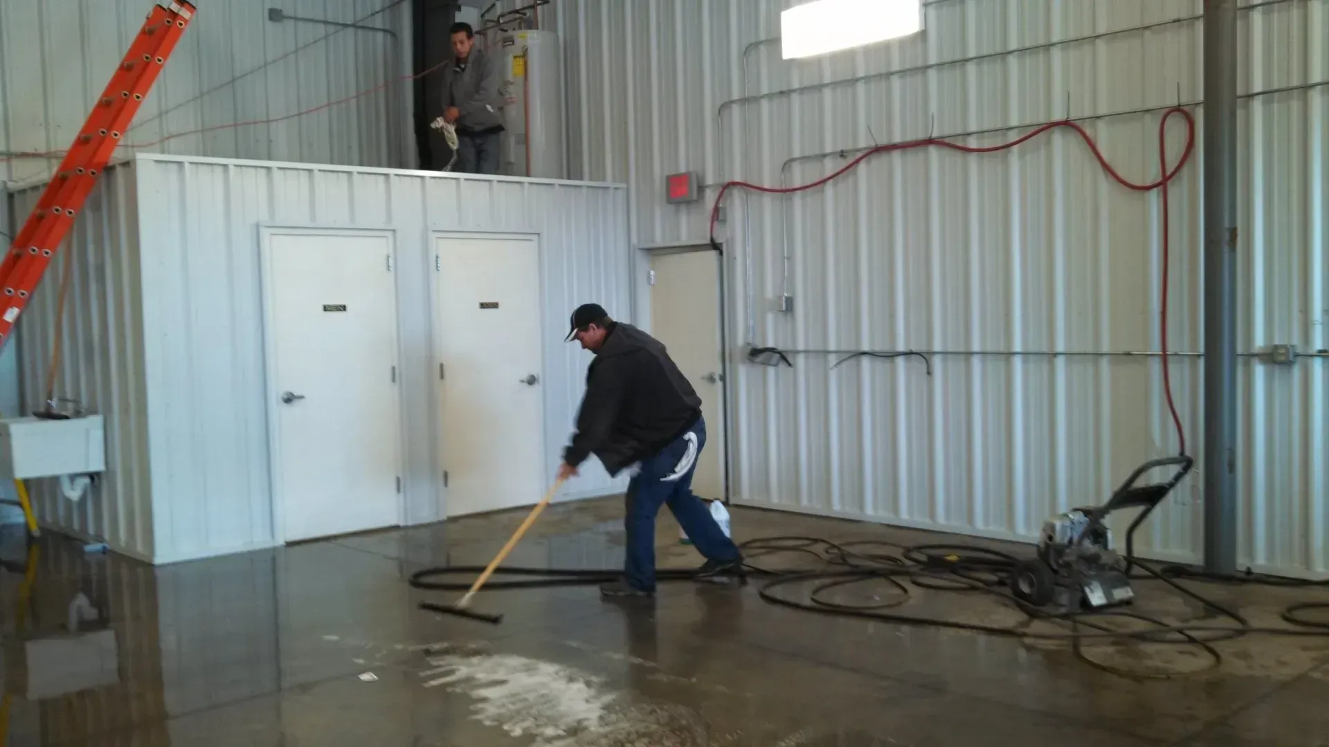 Two men cleaning a wet, gray-floored industrial space. One sweeps the floor, another works overhead.