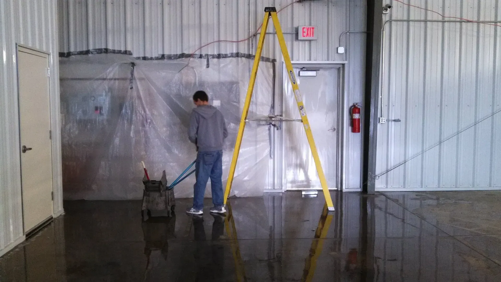 Person operating a floor cleaning machine in a warehouse. A-frame ladder, plastic sheeting, and a door are in the background.