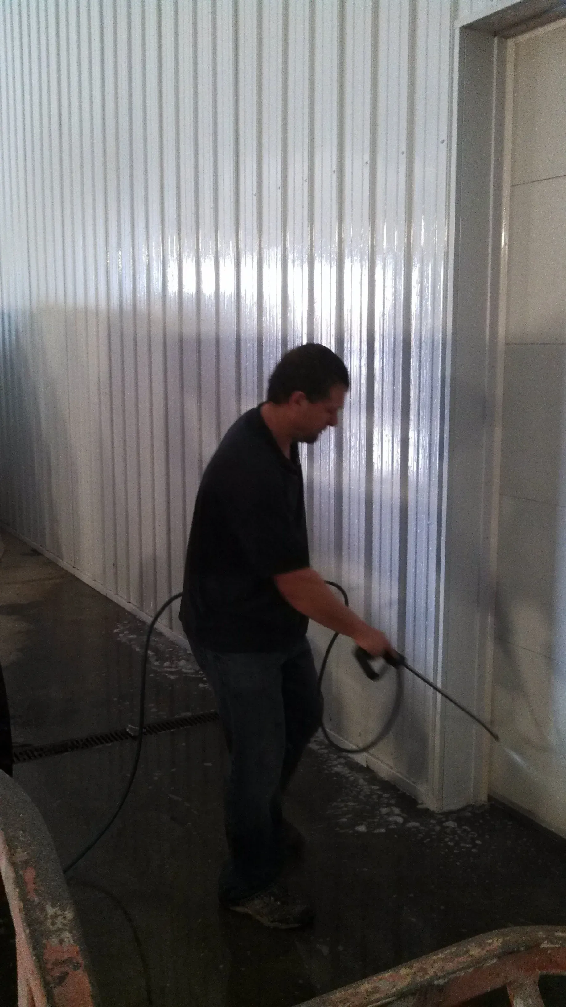 Man pressure washing a corrugated metal wall in a wet, concrete space.