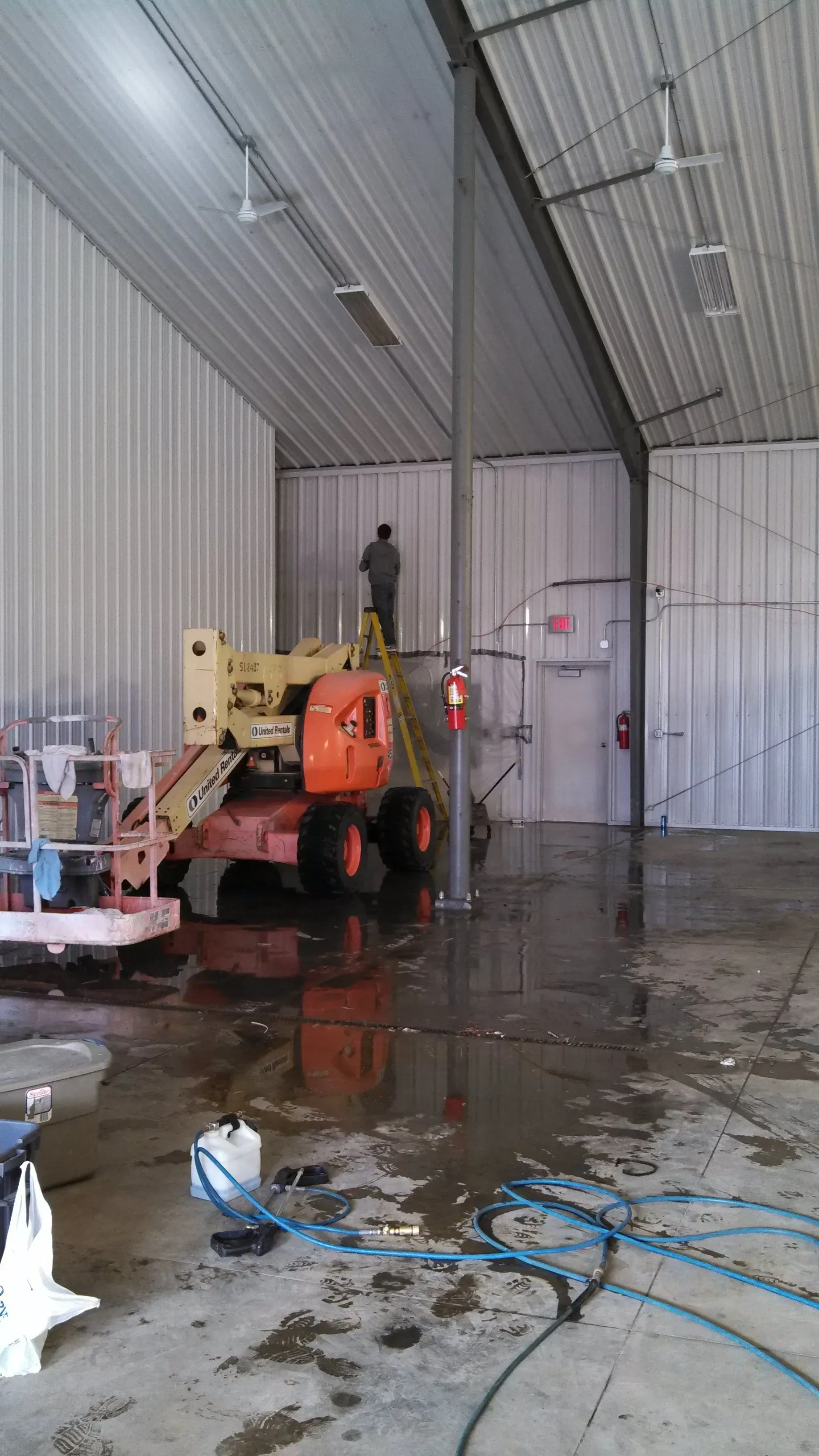Man on a ladder in a metal building, near an orange lift and wet floor.