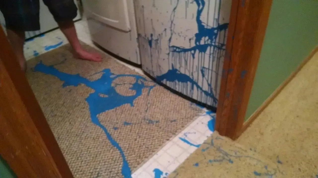 Blue paint splattered on carpet, walls, and appliance in a doorway. Bare foot visible.