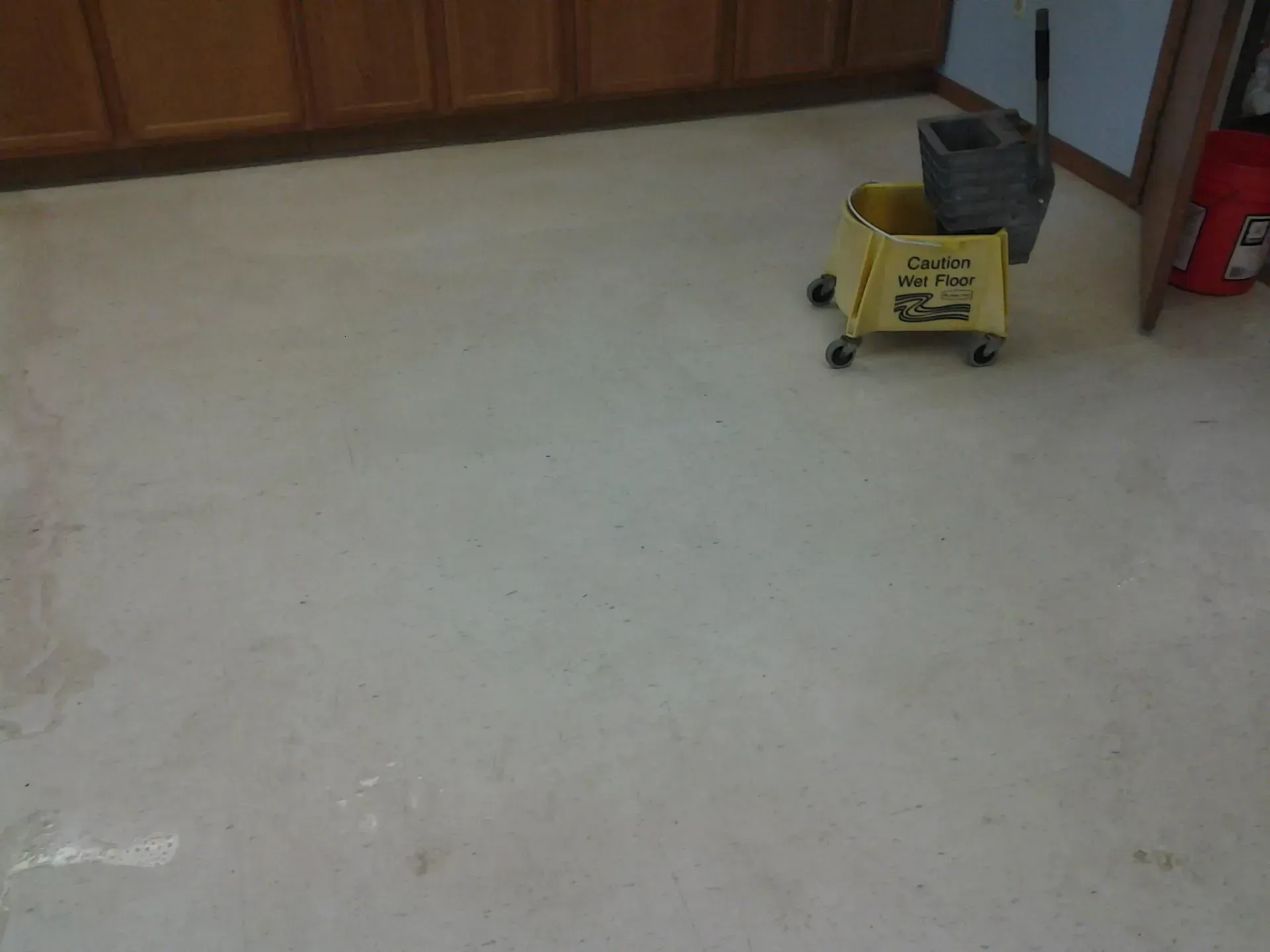 Off-white floor in a room. A yellow cleaning cart, a bucket, and a mop are present. Wooden cabinets are in the background.
