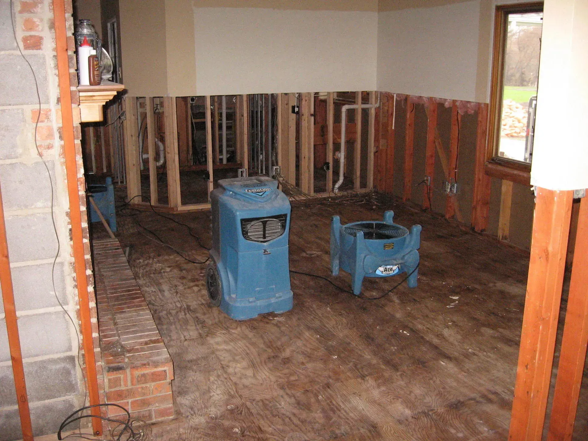 Room gutted due to water damage; wood framing exposed. Two blue dehumidifiers on wooden floor.