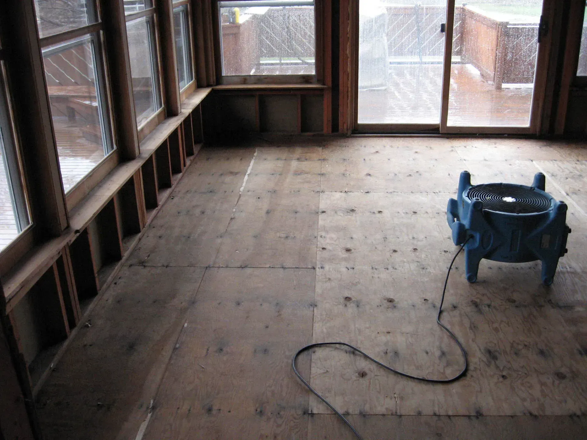 Interior of a room under construction, with exposed plywood floor, windows, and a blue water containment device.