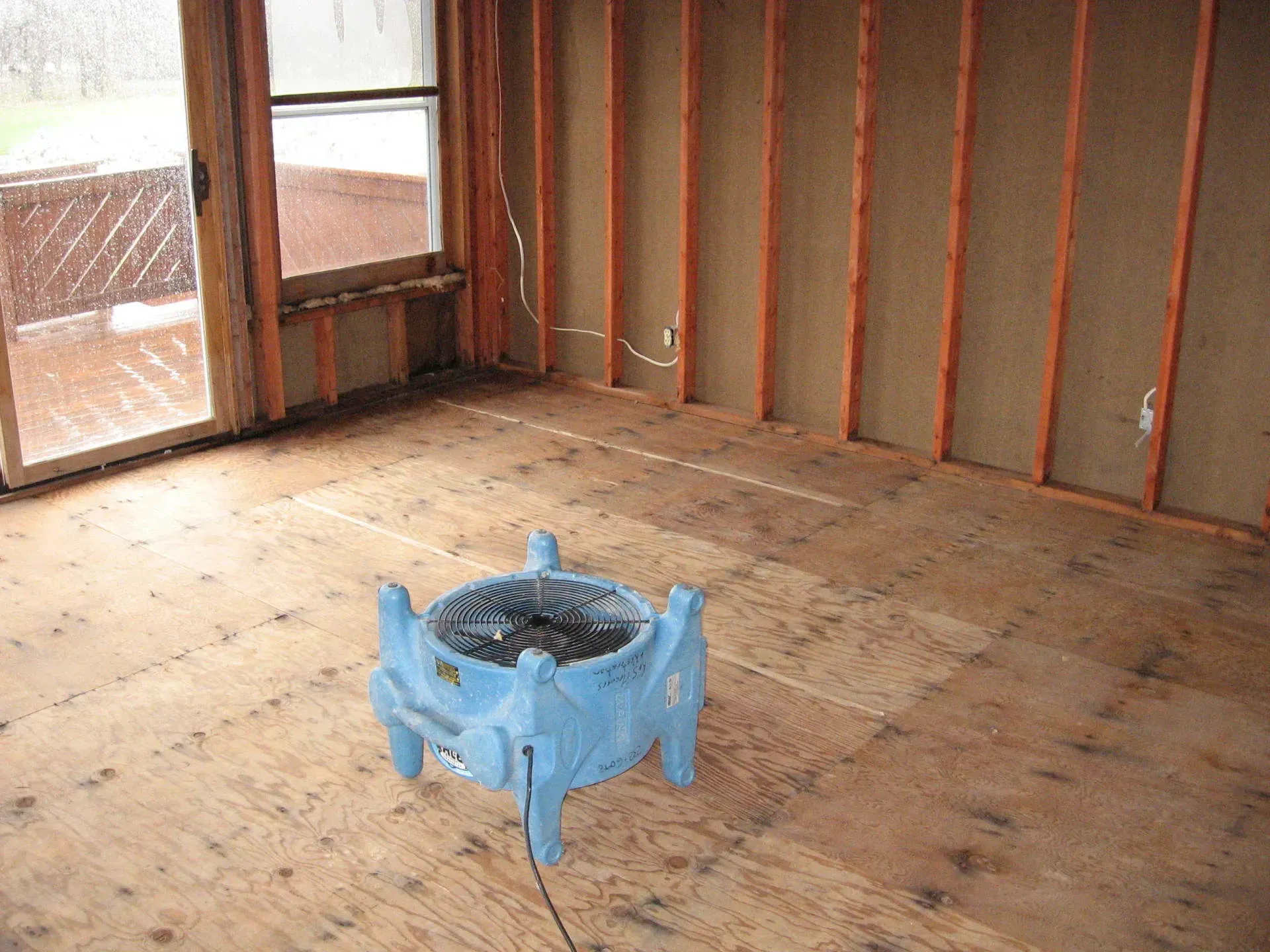 Blue air mover in a room with bare wood walls and flooring, near a sliding glass door.