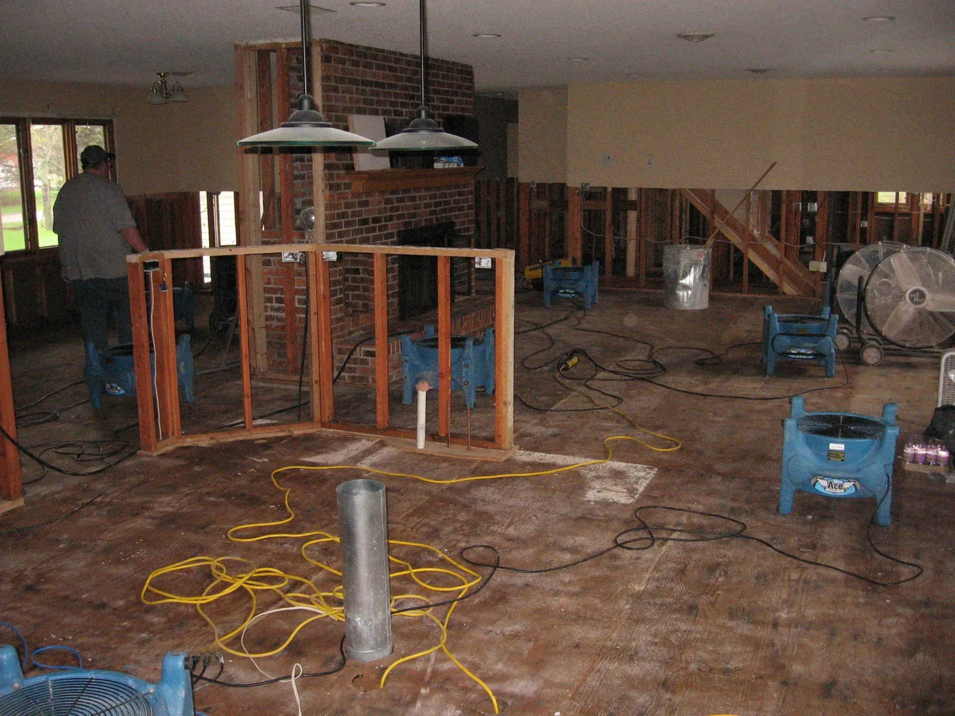 Room gutted after water damage, with framing exposed. Fans and hoses are drying the floor, a man stands near a wall.