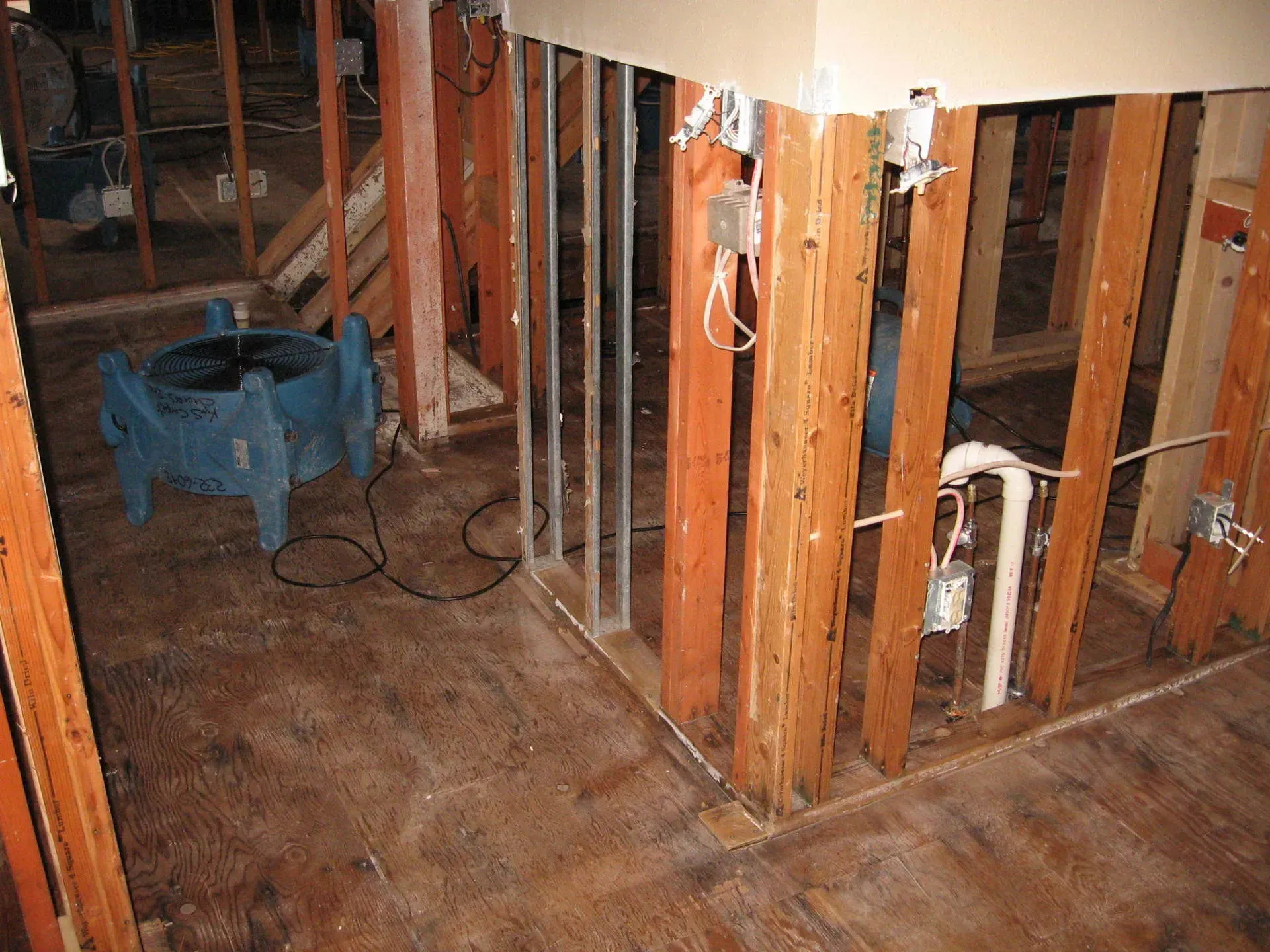 Interior view of a room under construction with exposed wooden beams, wiring, and a blue air mover.
