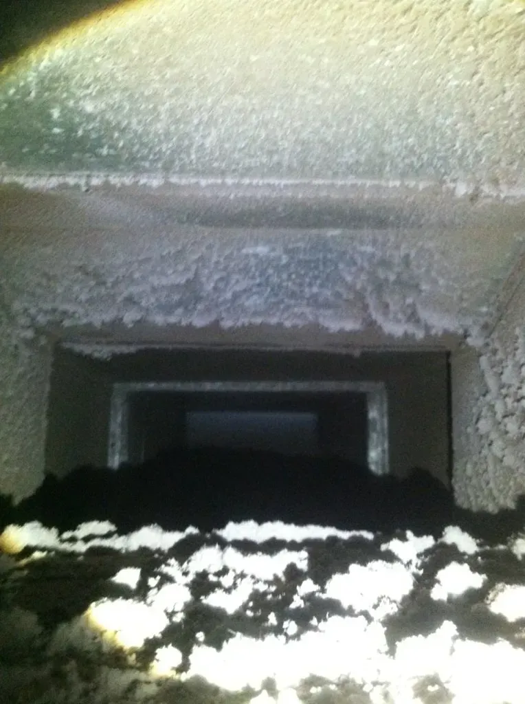 Inside of a dusty rectangular duct, filled with dark debris at the bottom and white substance along the sides.