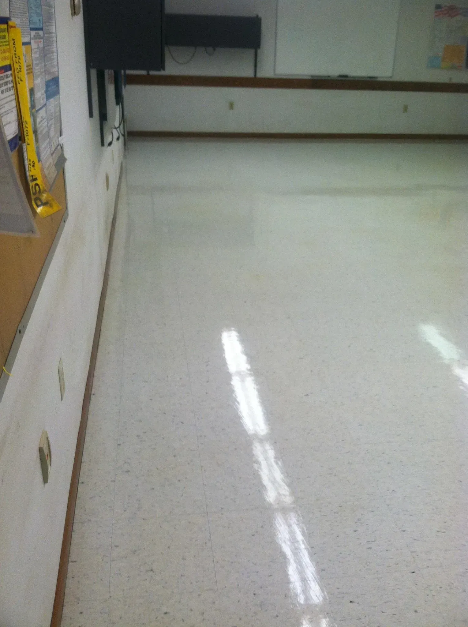 Light-colored, tiled floor with sunlight reflecting. A wall with a bulletin board and a counter are on the left.