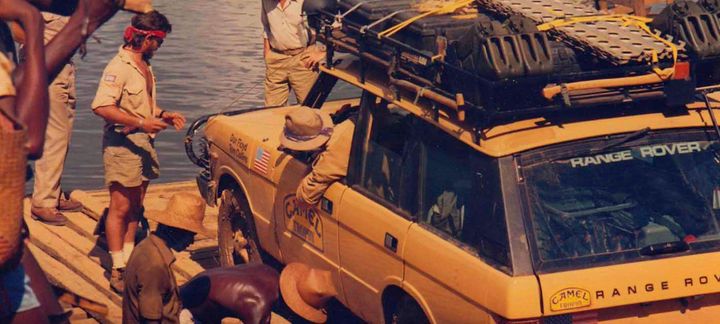 a man is standing next to a yellow range rover on a wooden raft .