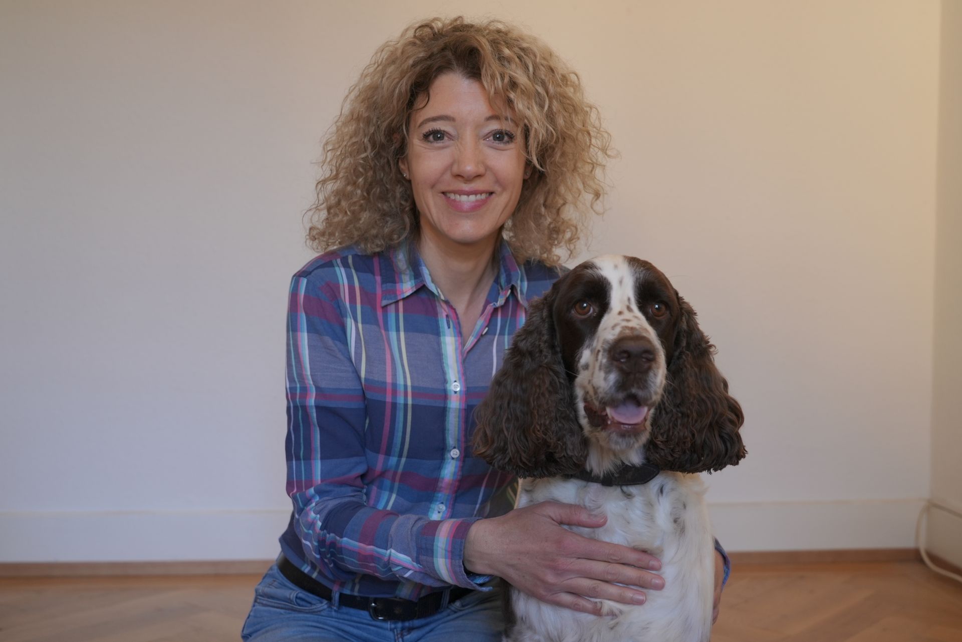 Une femme aux cheveux bouclés et son chien sourient. Ils se trouvent à l'intérieur, près d'un mur clair et d'un parquet.