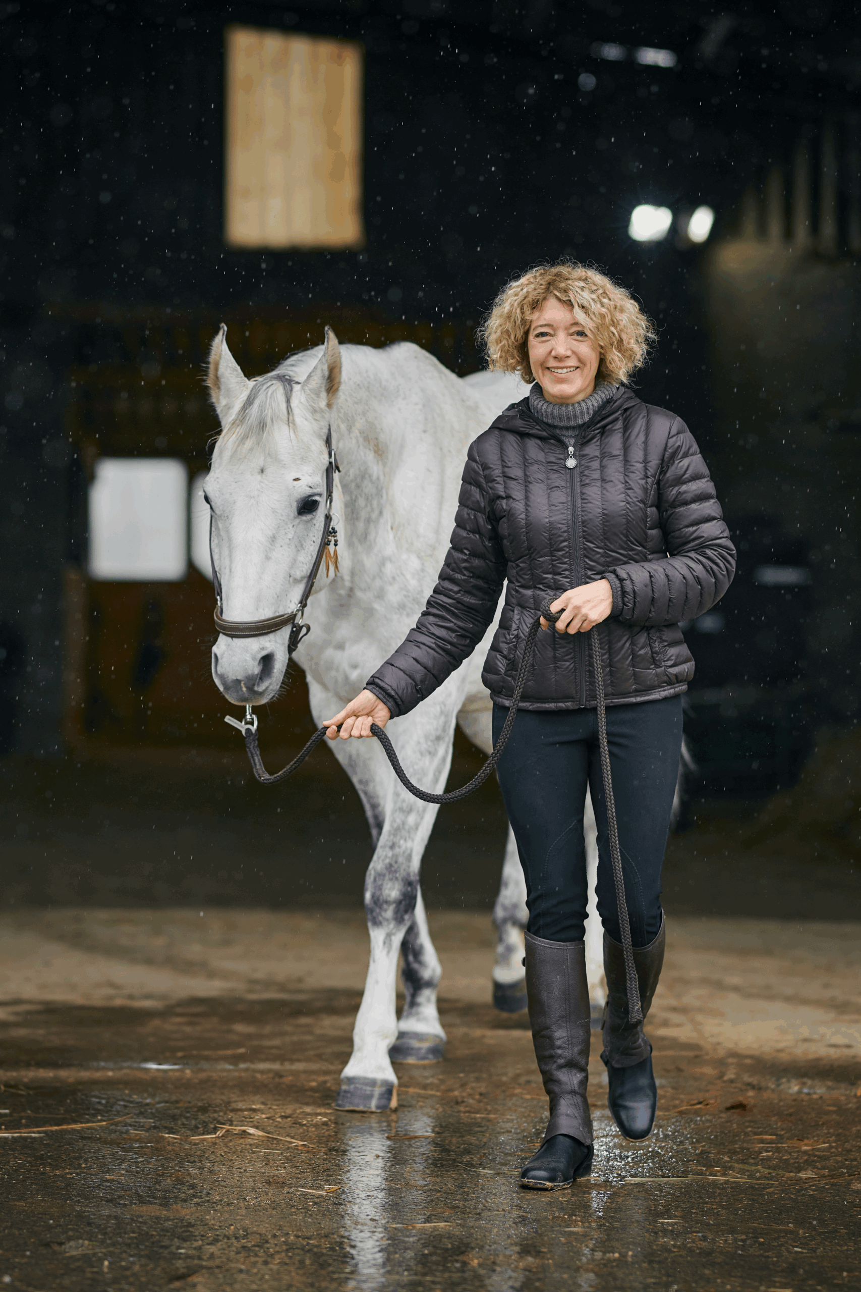Une femme mène un cheval gris hors de l'écurie, en souriant. Il pleut.