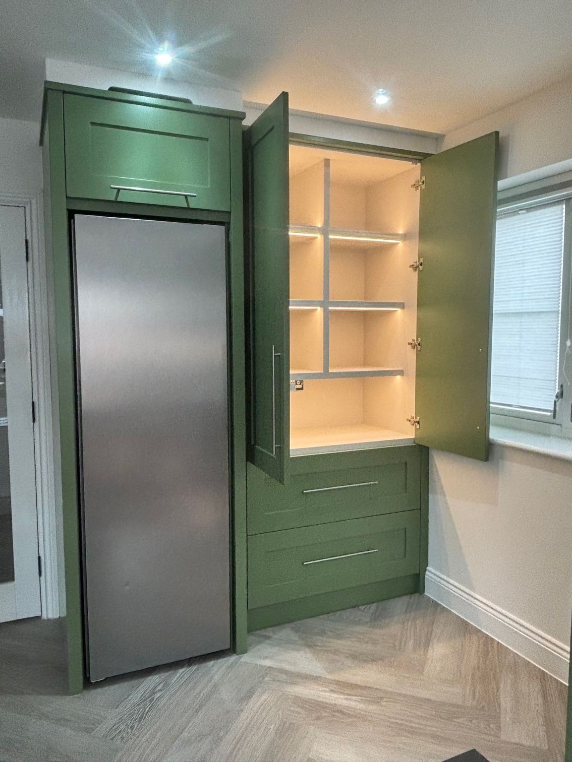 Green kitchen cabinetry with a stainless steel refrigerator and pantry, open to reveal lit shelves.