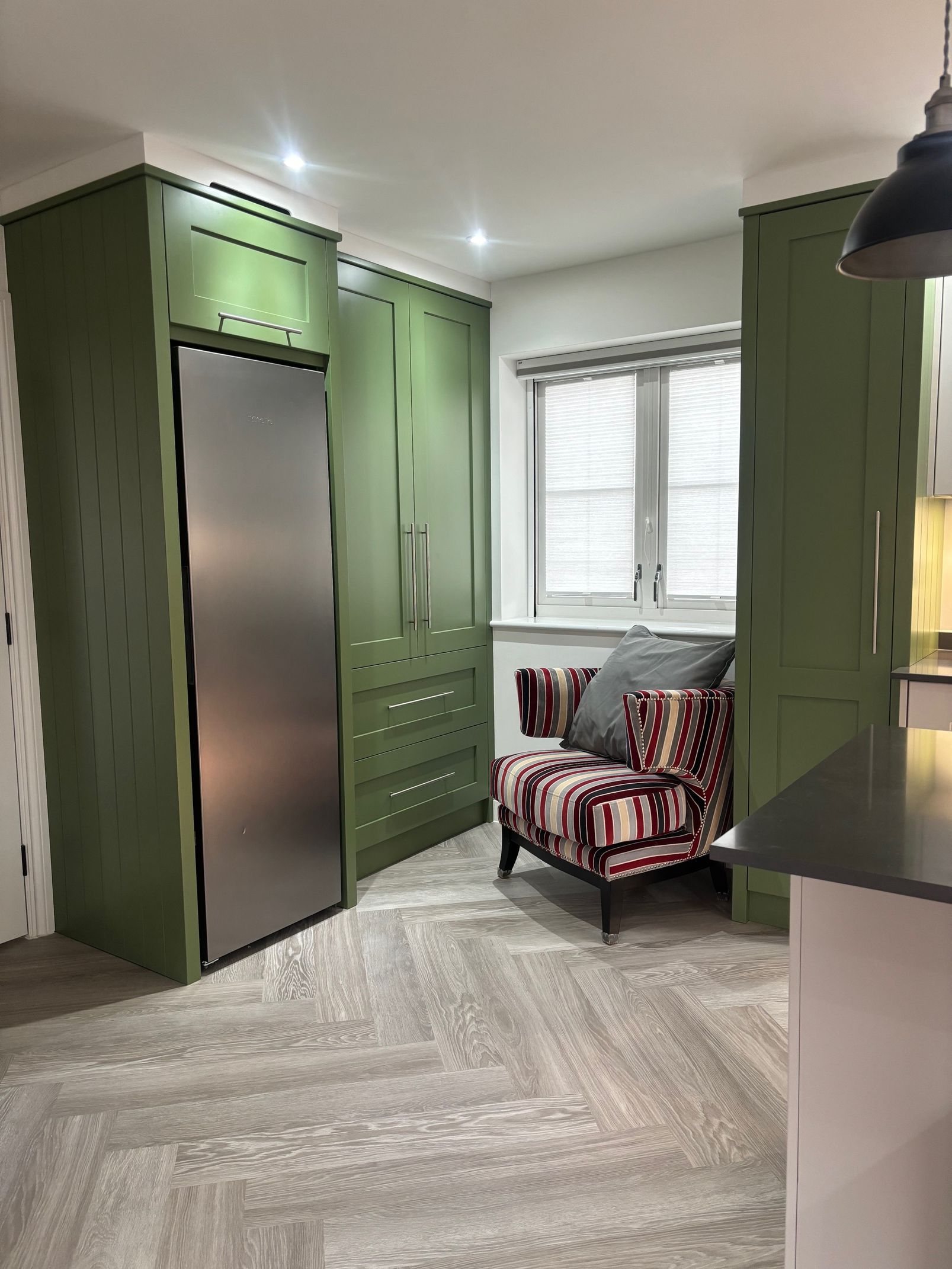 Green kitchen cabinets with stainless steel refrigerator, patterned chair by window.